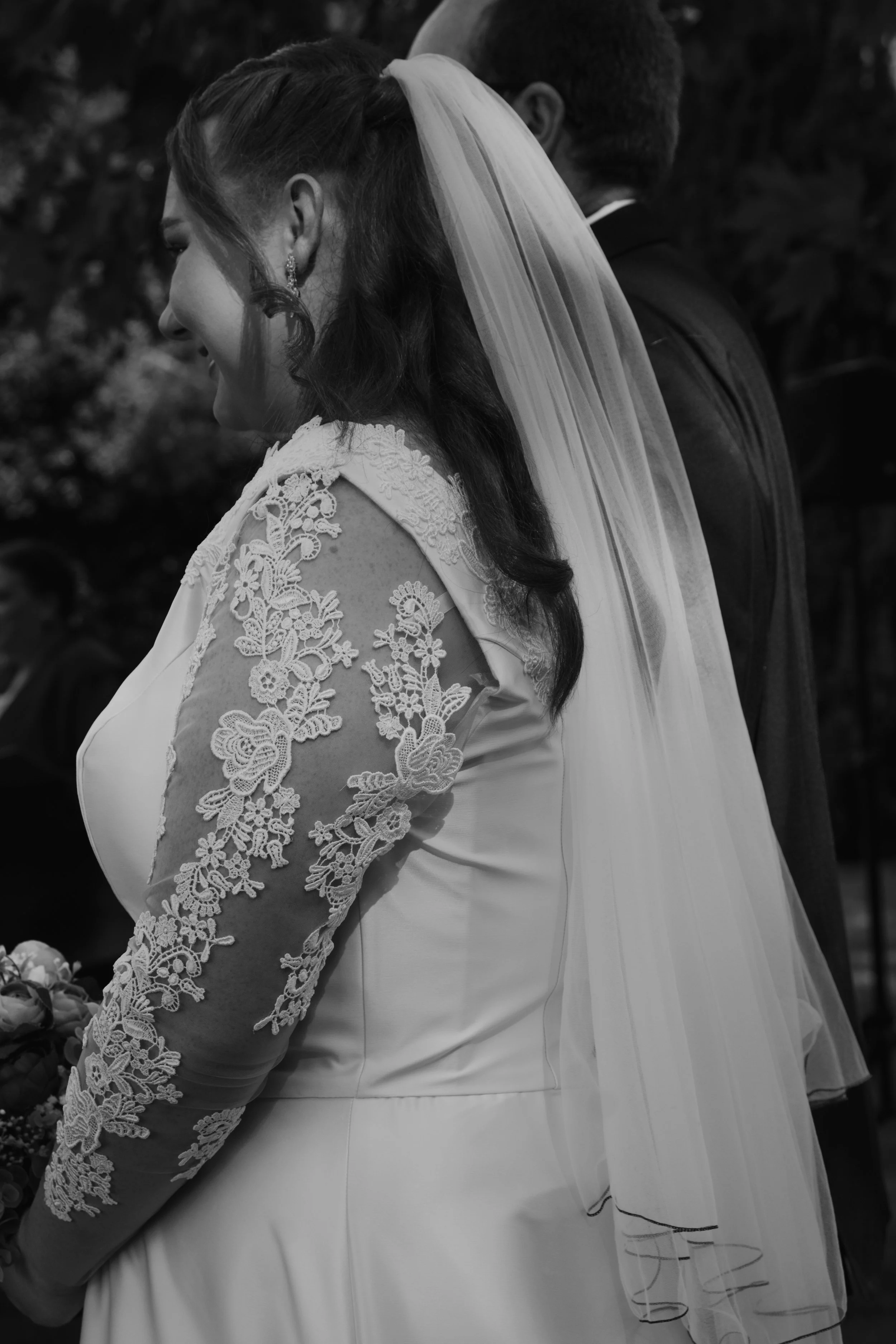 A bride in a wedding dress with lace sleeves and a veil, smiling in a moment of ceremony, with a groom in a suit standing beside her.
