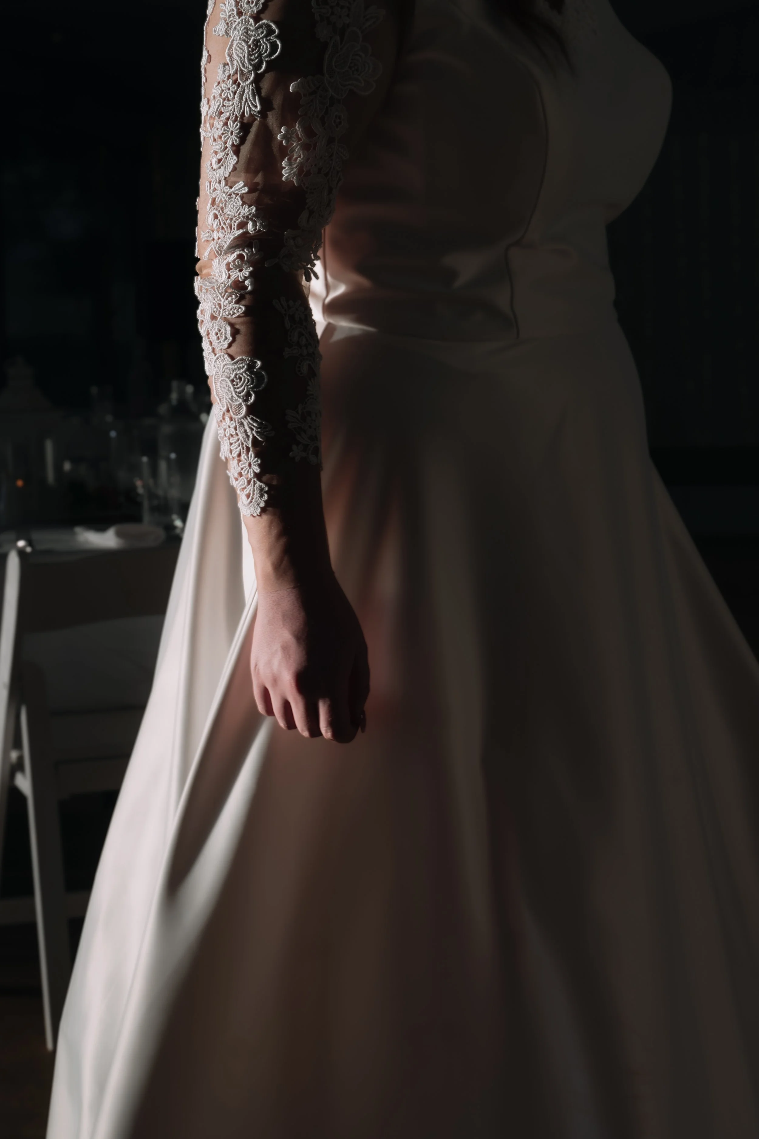 Close-up of a bride in a white wedding gown with lace sleeves, standing in a dimly lit room.