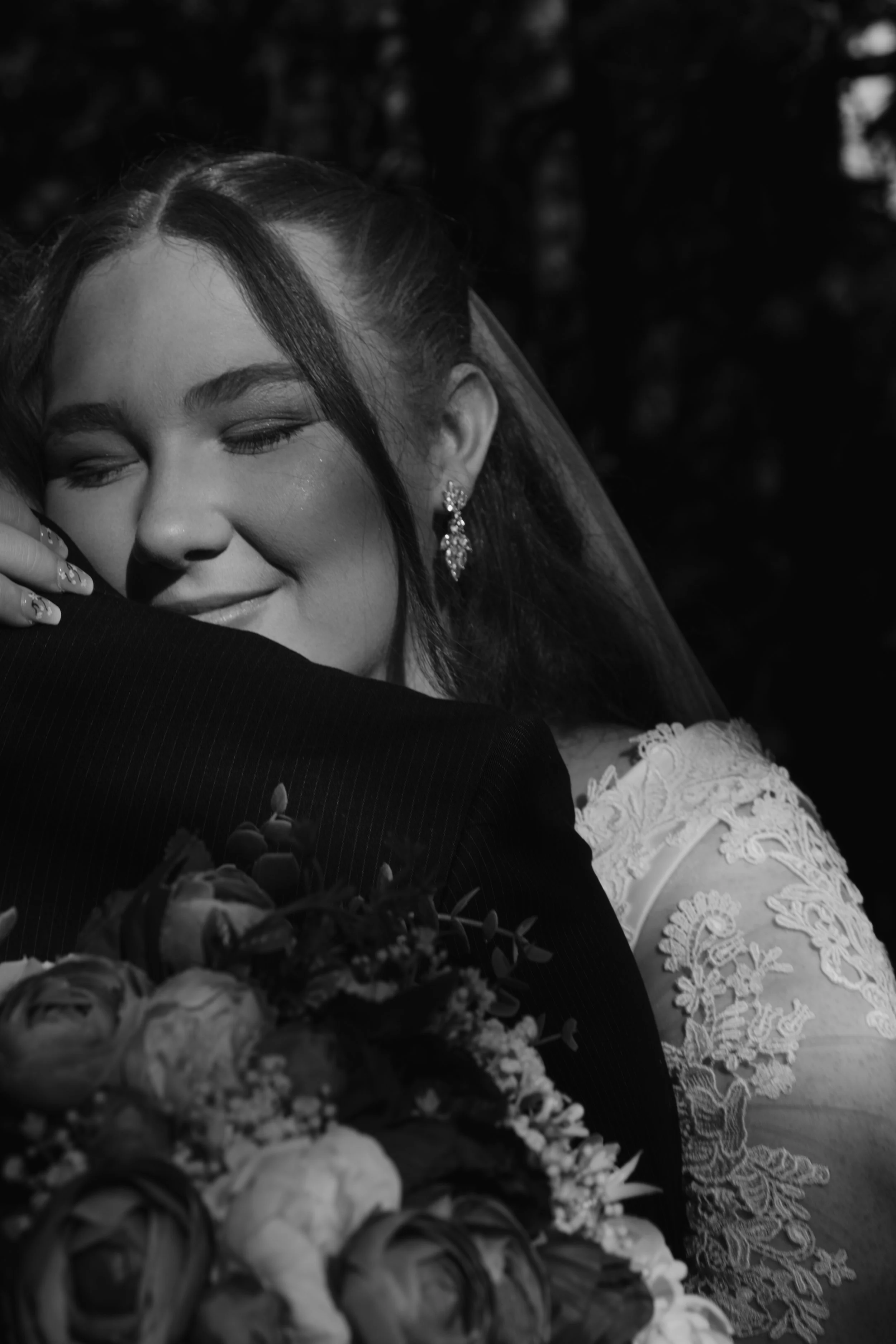 A black and white photo of a woman with long dark hair and earrings, smiling with her eyes closed, hugging someone in formal attire, likely during a wedding or special event.