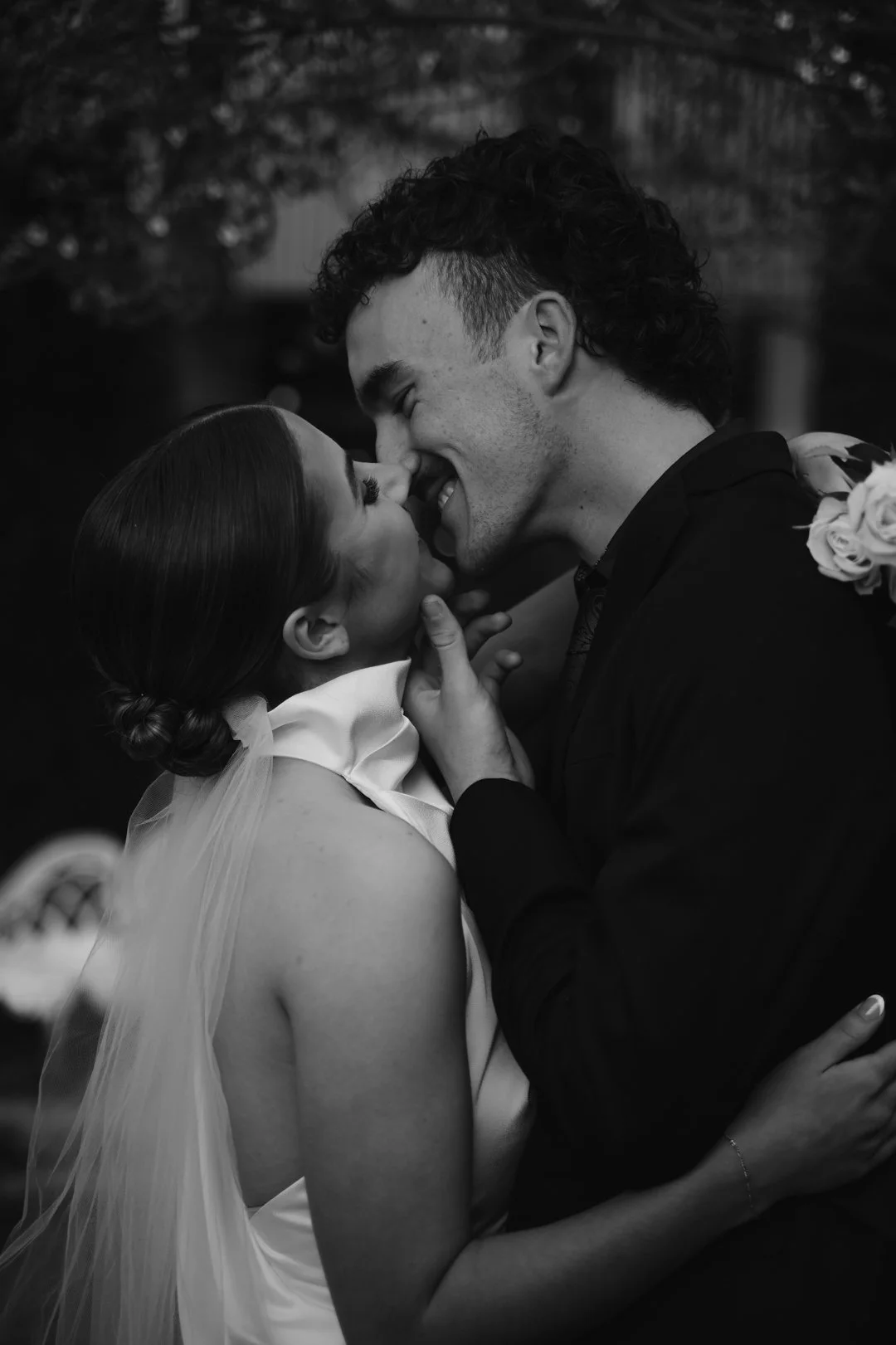 A black-and-white photo of a couple sharing a kiss, with the woman in a wedding gown and the man in a dark suit, both smiling and close together.