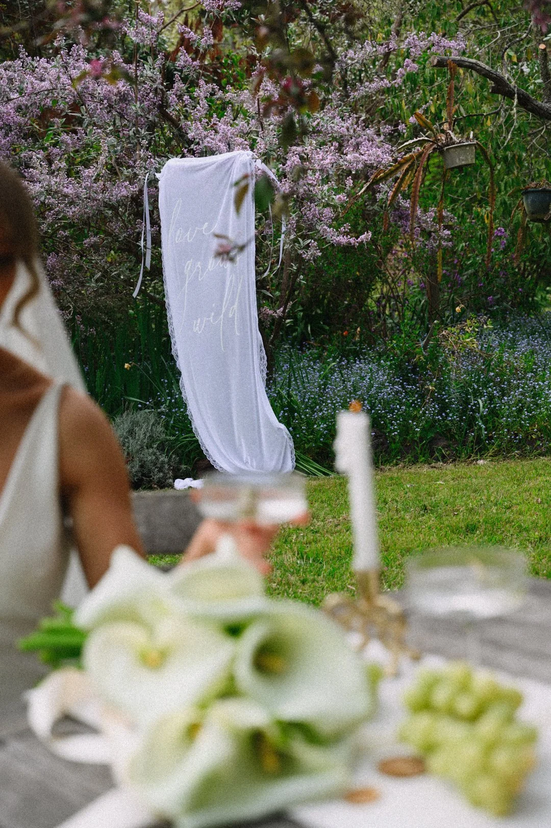 A garden with pink and purple flowering shrubs and greenery, featuring a white fabric with handwritten words 'love, peace, wild' hanging among the plants.