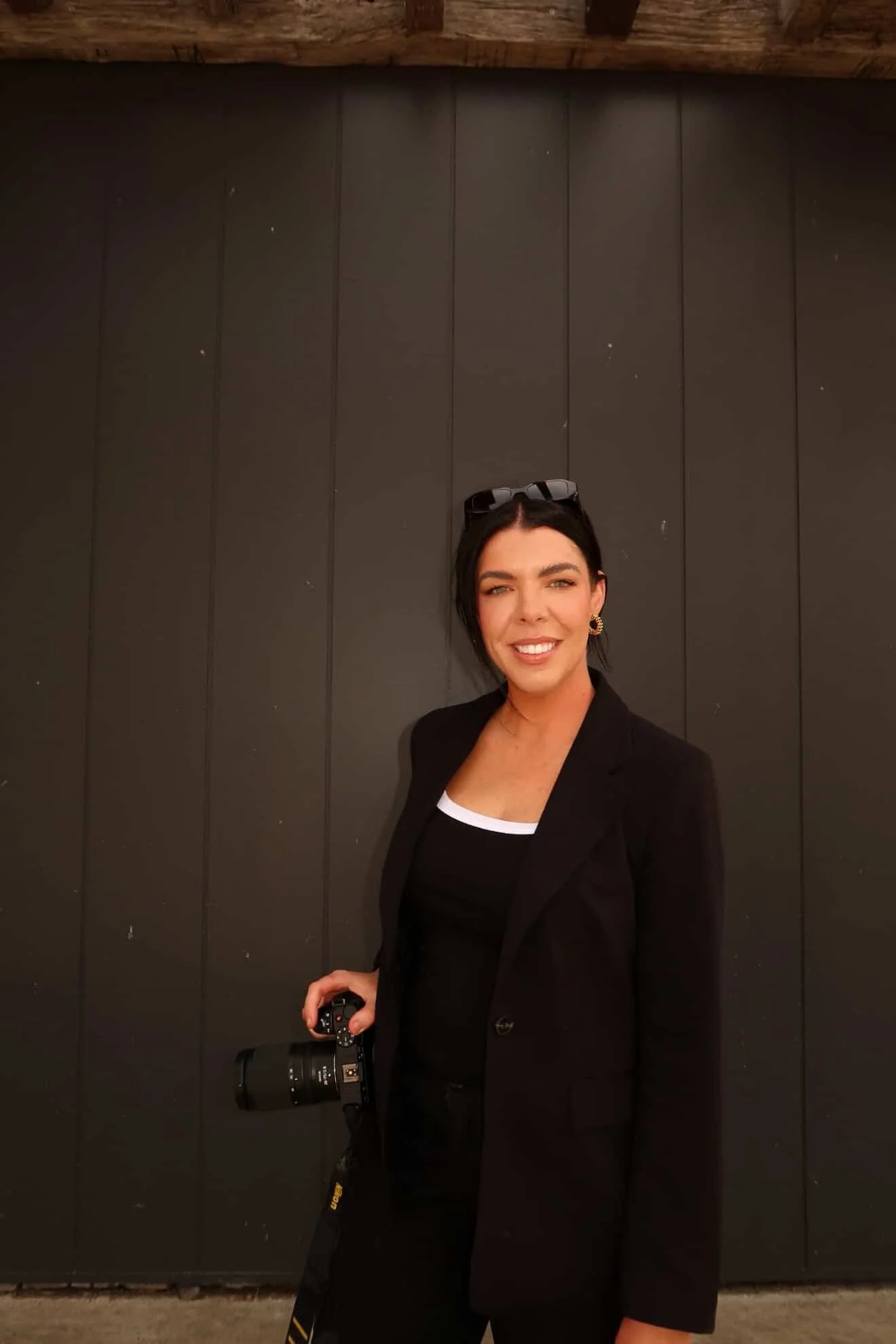 A woman smiling, wearing a black blazer and top with a white neckline trim, black sunglasses on her head, and gold hoop earrings, holding a camera with a zoom lens, standing against a black paneled wall.