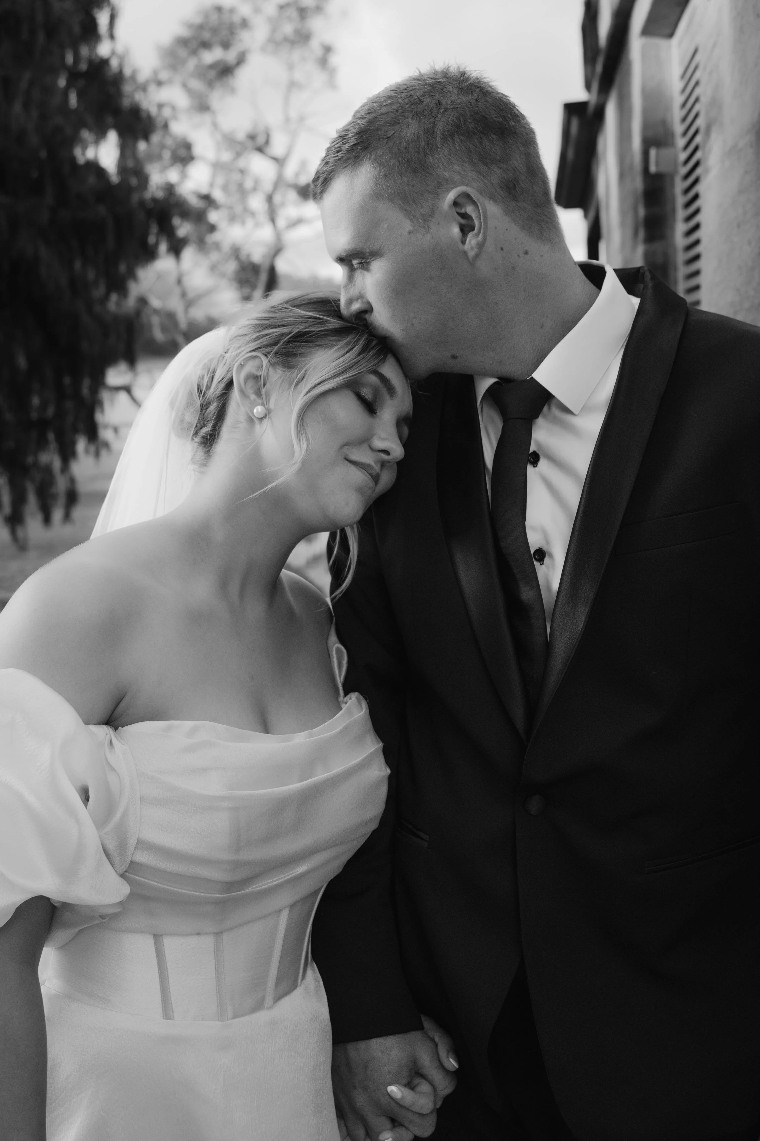 A black-and-white photograph of a newlywed couple holding hands, with the groom kissing the bride's forehead. The bride is wearing an off-shoulder wedding dress and the groom is in a tuxedo, standing outdoors near a building with trees in the background.