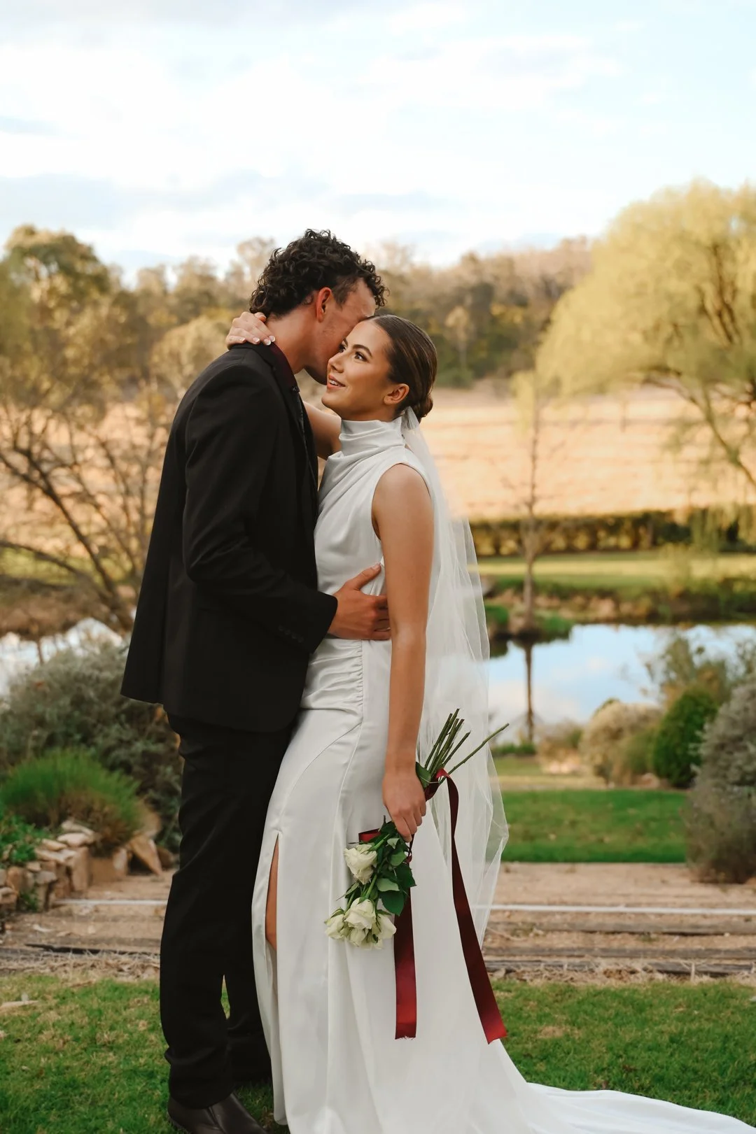 A newlywed couple stands close together outdoors by a pond and trees, with the bride holding a bouquet of white roses and the groom gently touching her.