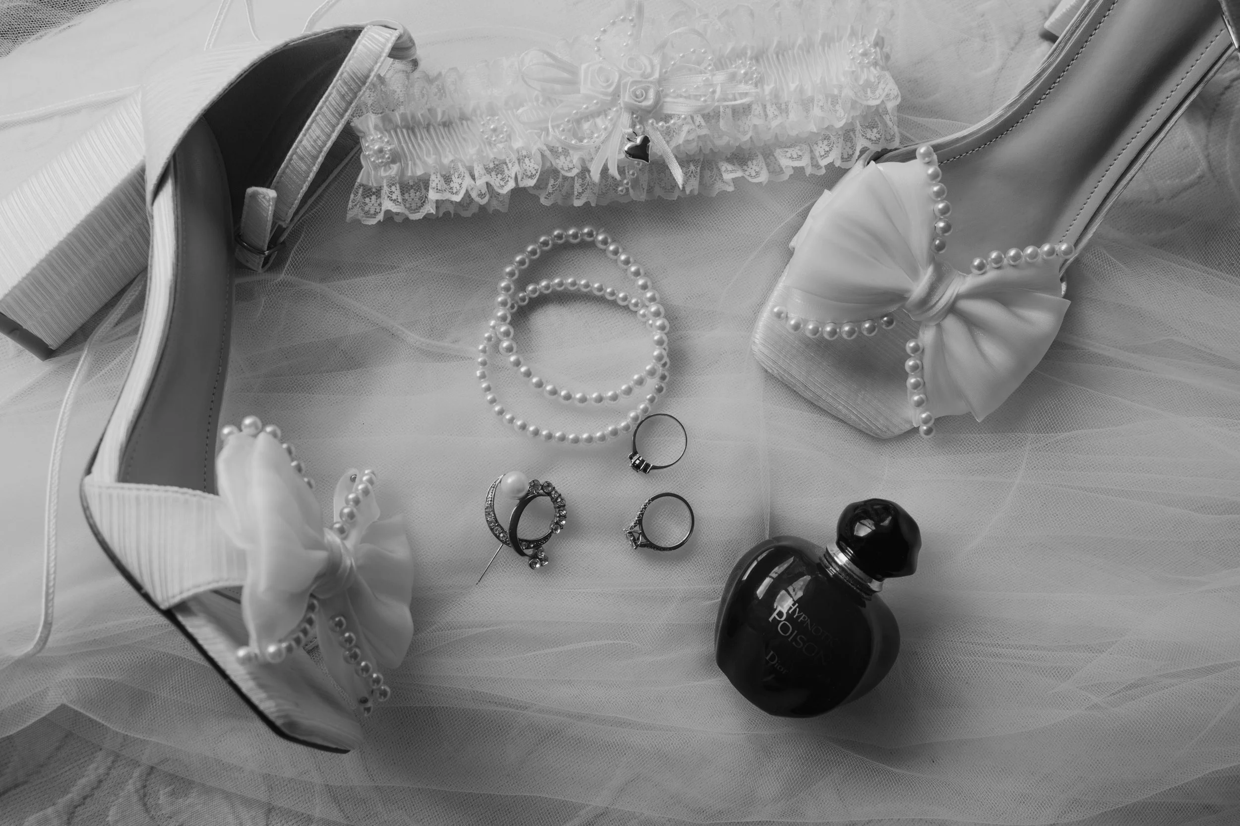 Arrangement of bridal accessories, including pearl-studded high heels with bows, pearl necklaces, rings, and a black perfume bottle, on a soft fabric surface.
