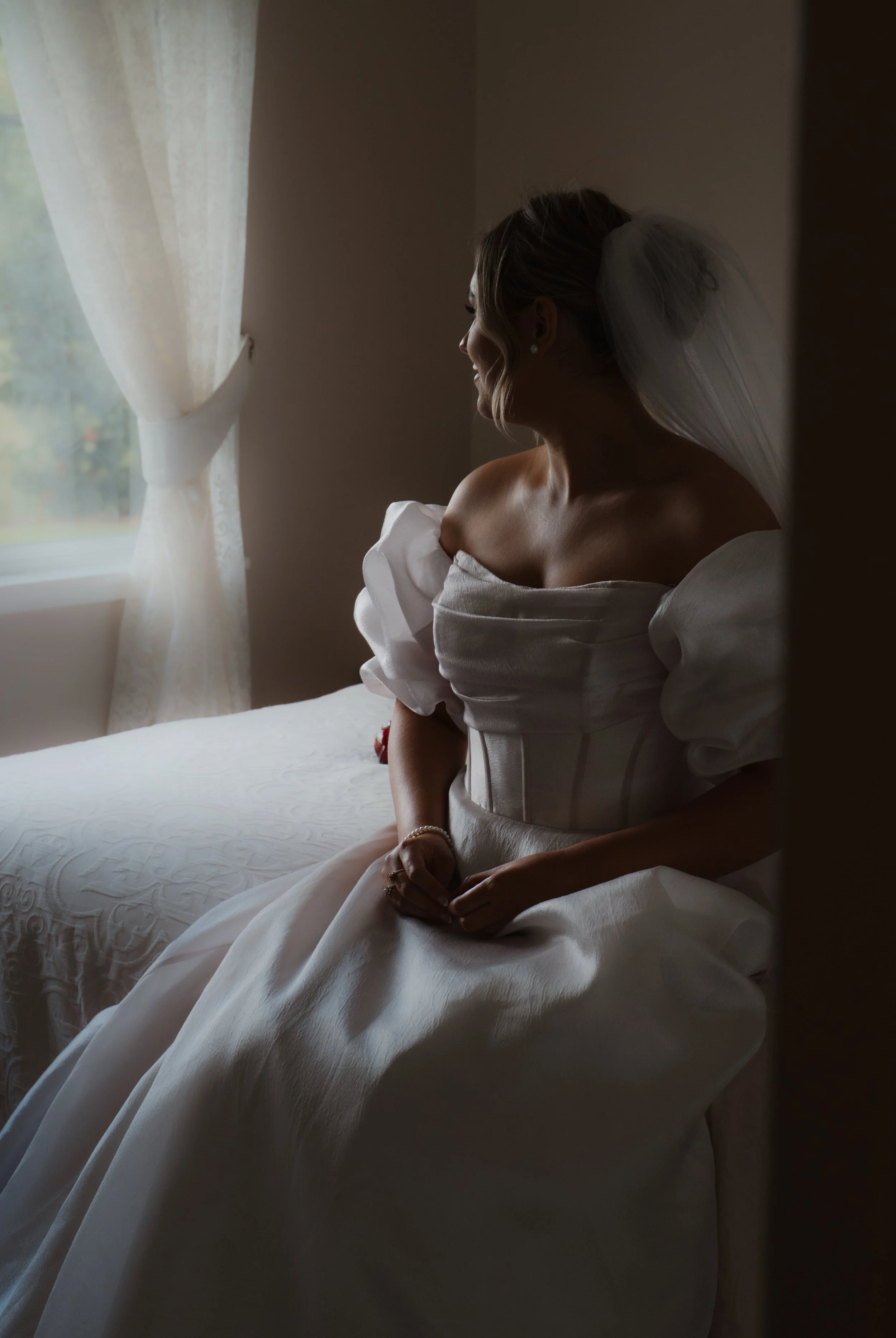 A bride sitting on a bed near a window with white curtains, looking out at the light.
