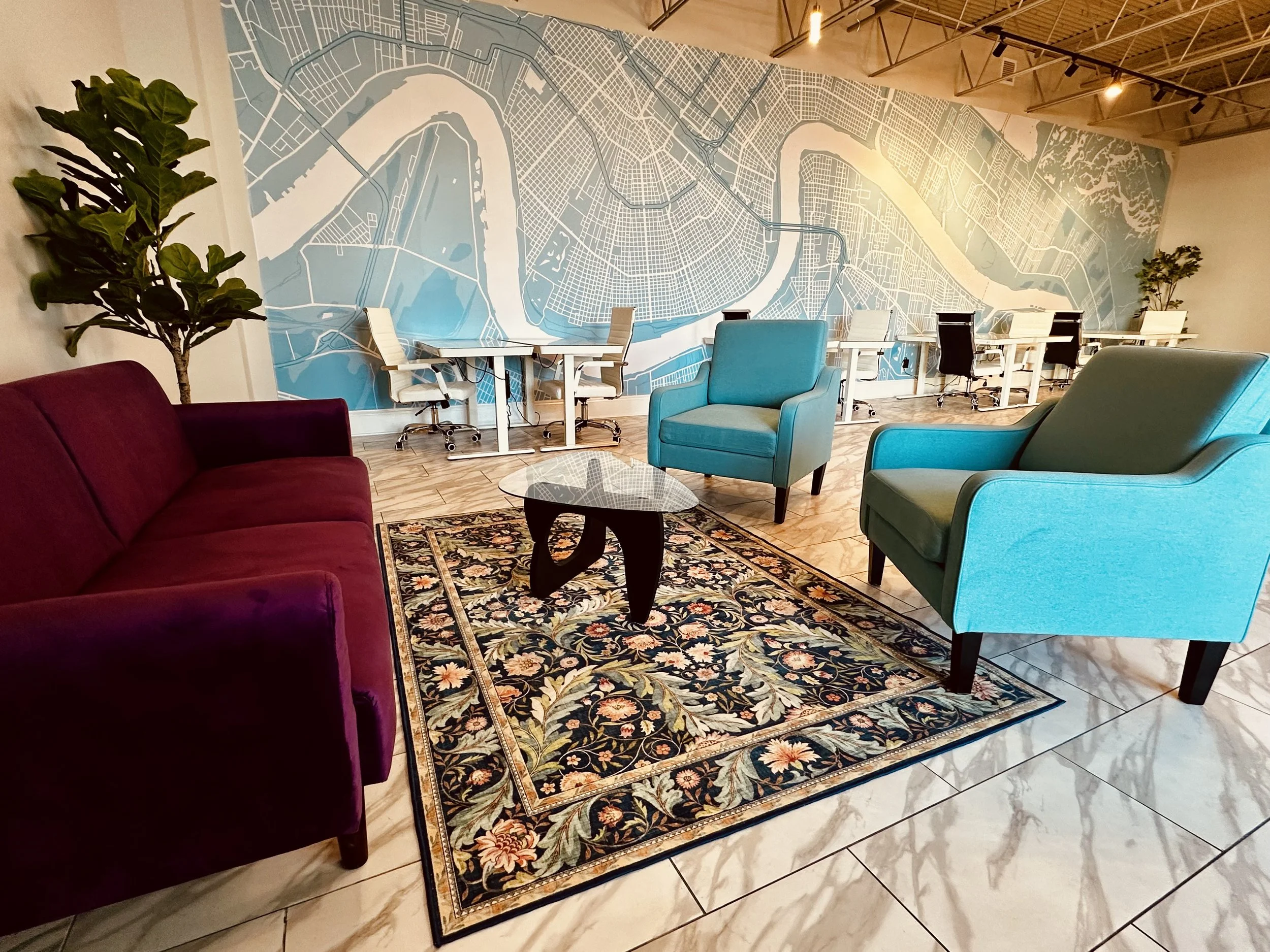 A modern office lounge area with a vintage floral area rug, a maroon sofa, two turquoise armchairs, a glass-top coffee table, a large city map mural on the wall, a potted plant near the sofa, and a row of desks with white chairs and a city map mural in the background.