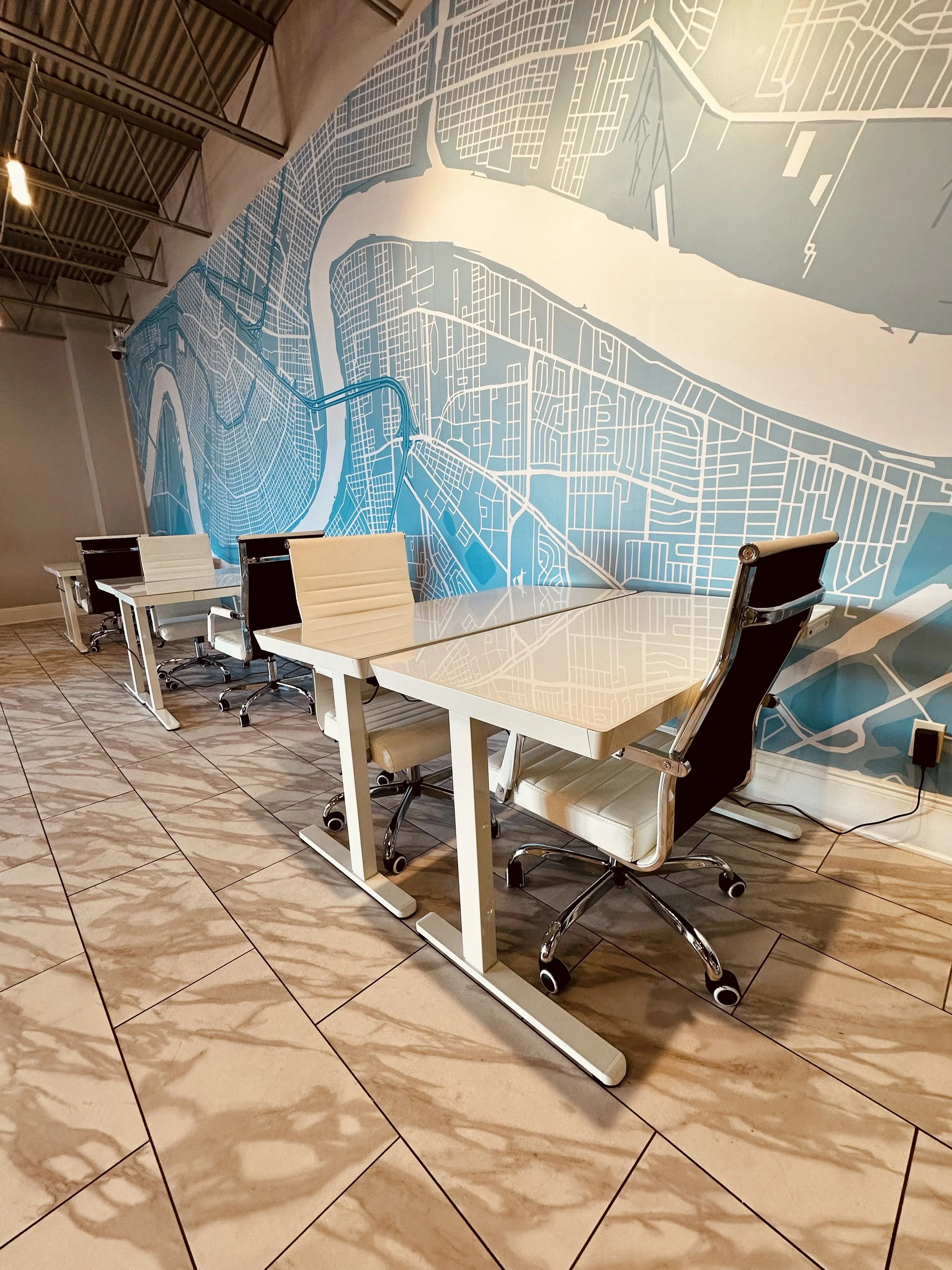 A modern office conference room with a large white table and several white office chairs with black backs. The wall features a large map or city layout in blue and white, and the floor has beige marble tiles. There is an electrical outlet visible on the wall, with a black cord plugged into it.