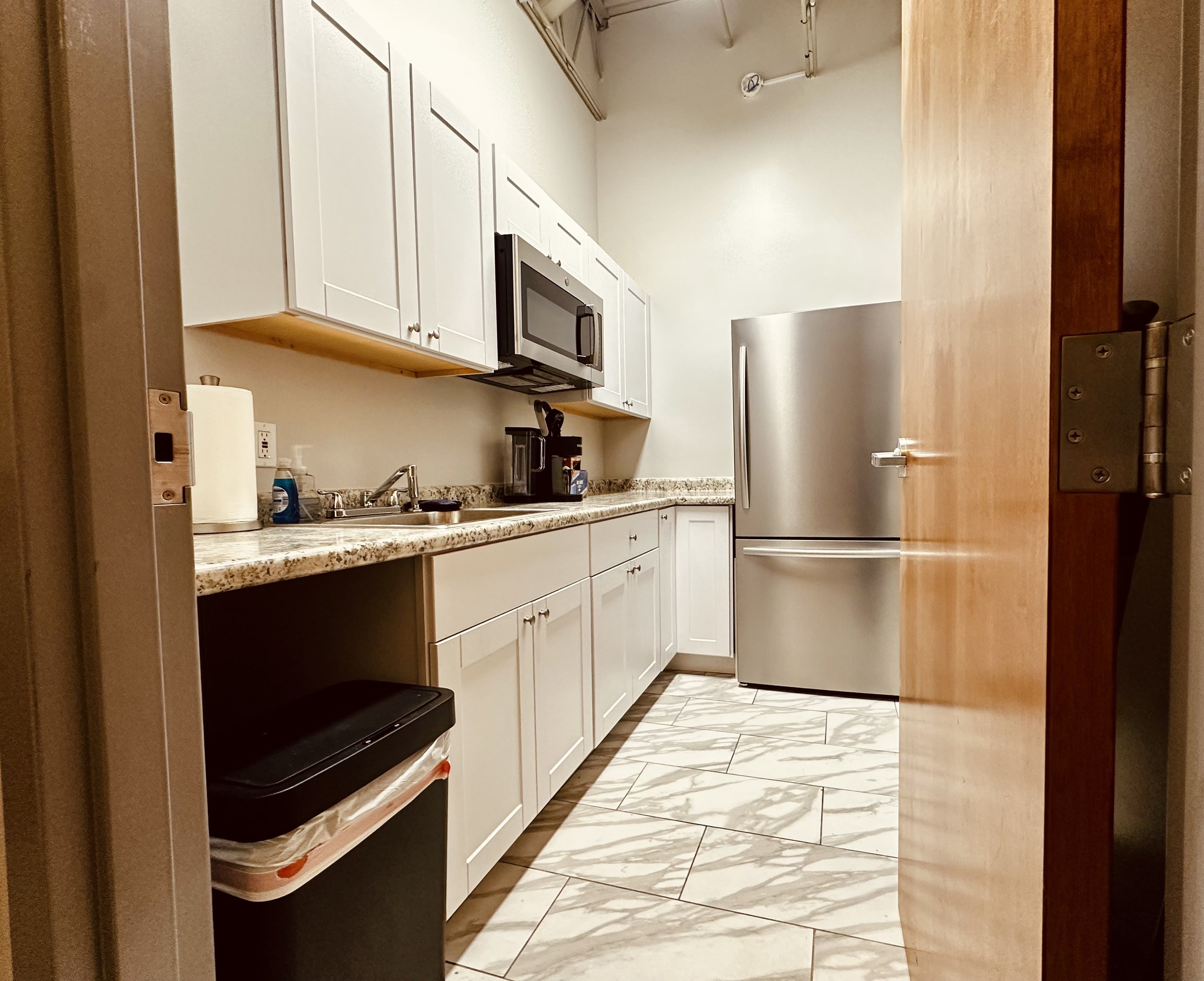 A small kitchen with white cabinets, a granite countertop, a stainless steel refrigerator, a microwave, and a coffee maker. The floor has white tiles with gray veins, and a wooden door is partially open.