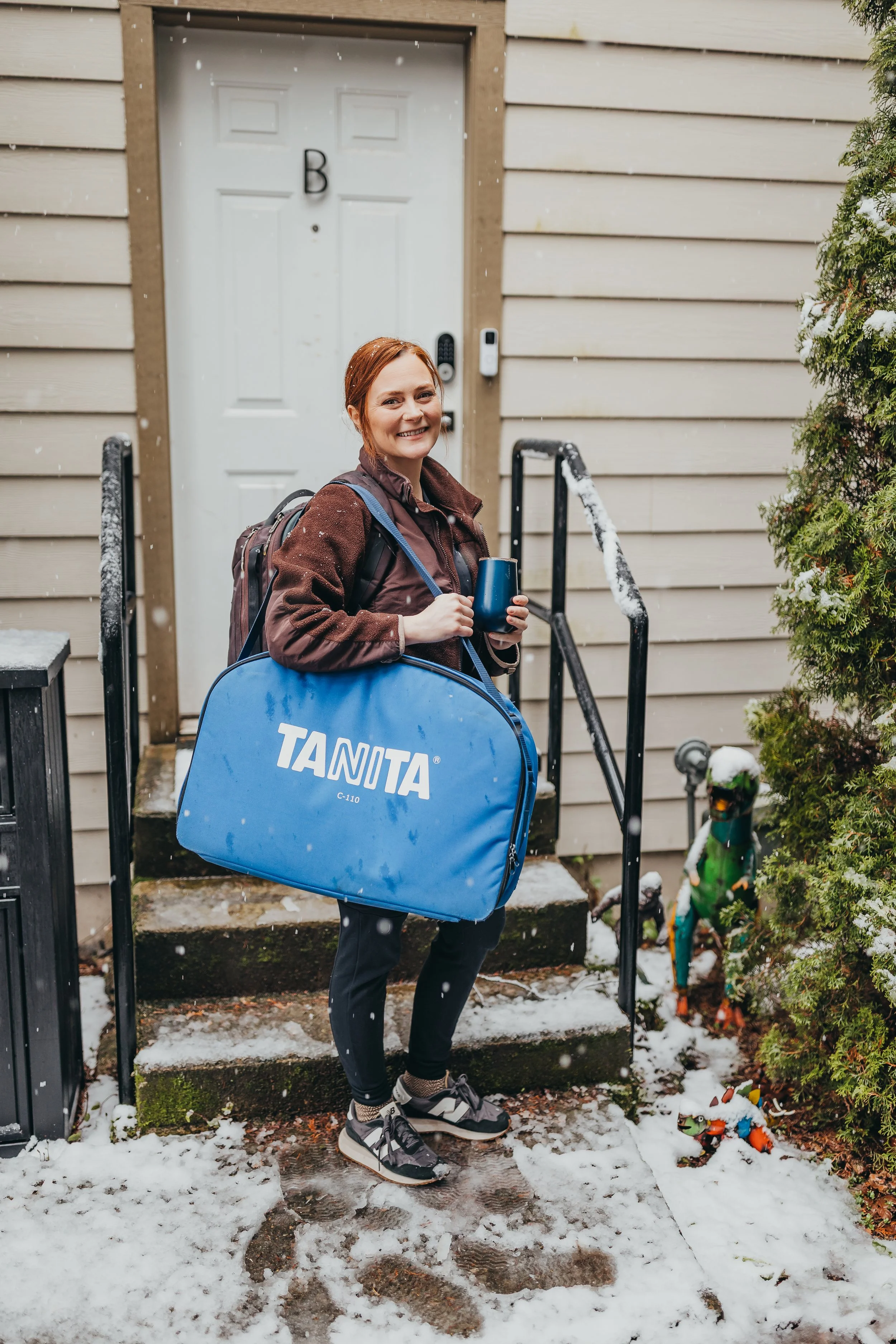 A woman, lying on a couch, is breastfeeding her newborn while a caregiver lactation consultant, wearing blue gloves, assists and observes, in a bright room with a window in the background.