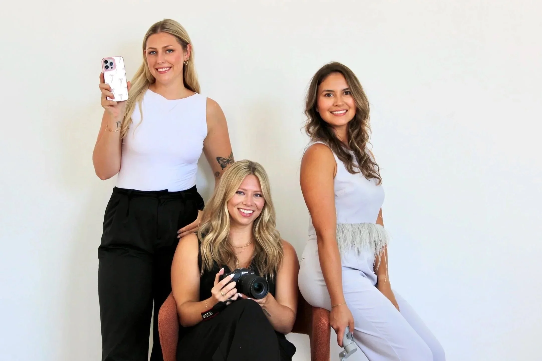 Three women smiling, two holding cameras or smartphones, against a plain white background.