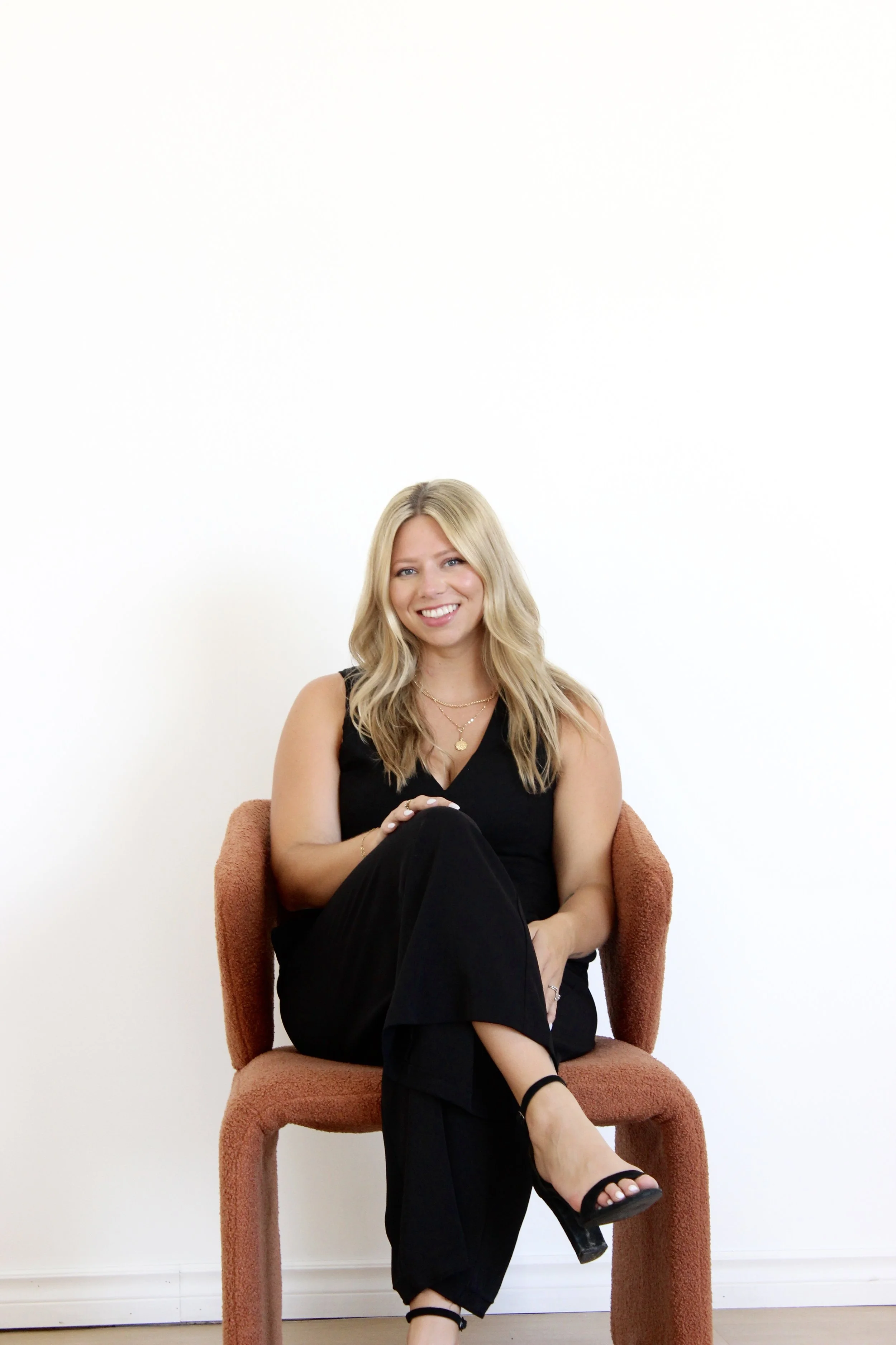 A woman with blonde hair sitting on a burnt orange chair, smiling, wearing a black sleeveless outfit and black high heels, with a white wall in the background.