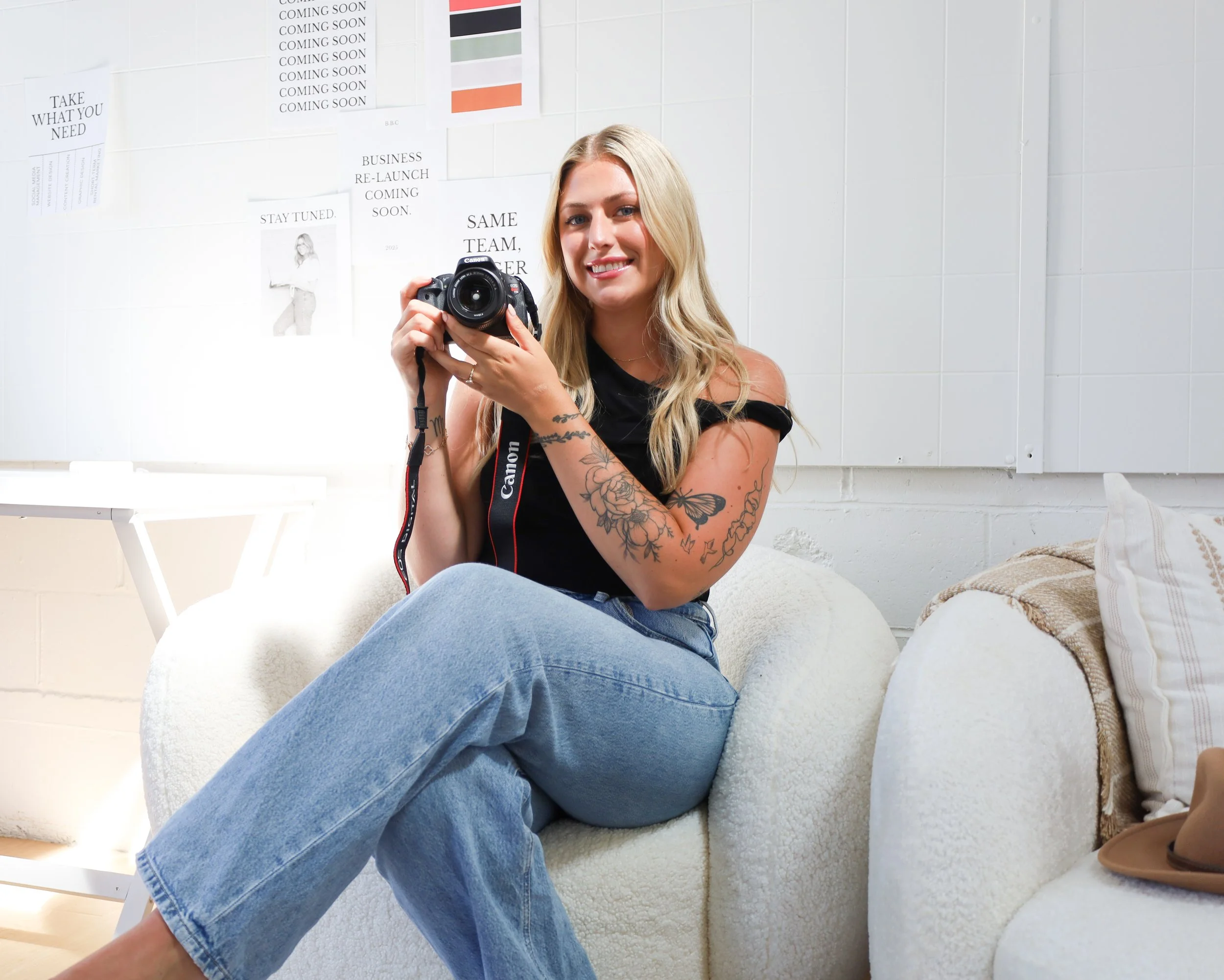 A smiling woman with long blonde hair and tattoos, sitting on a white comfy chair, holding a Canon camera, in a room with white brick walls and various posters.