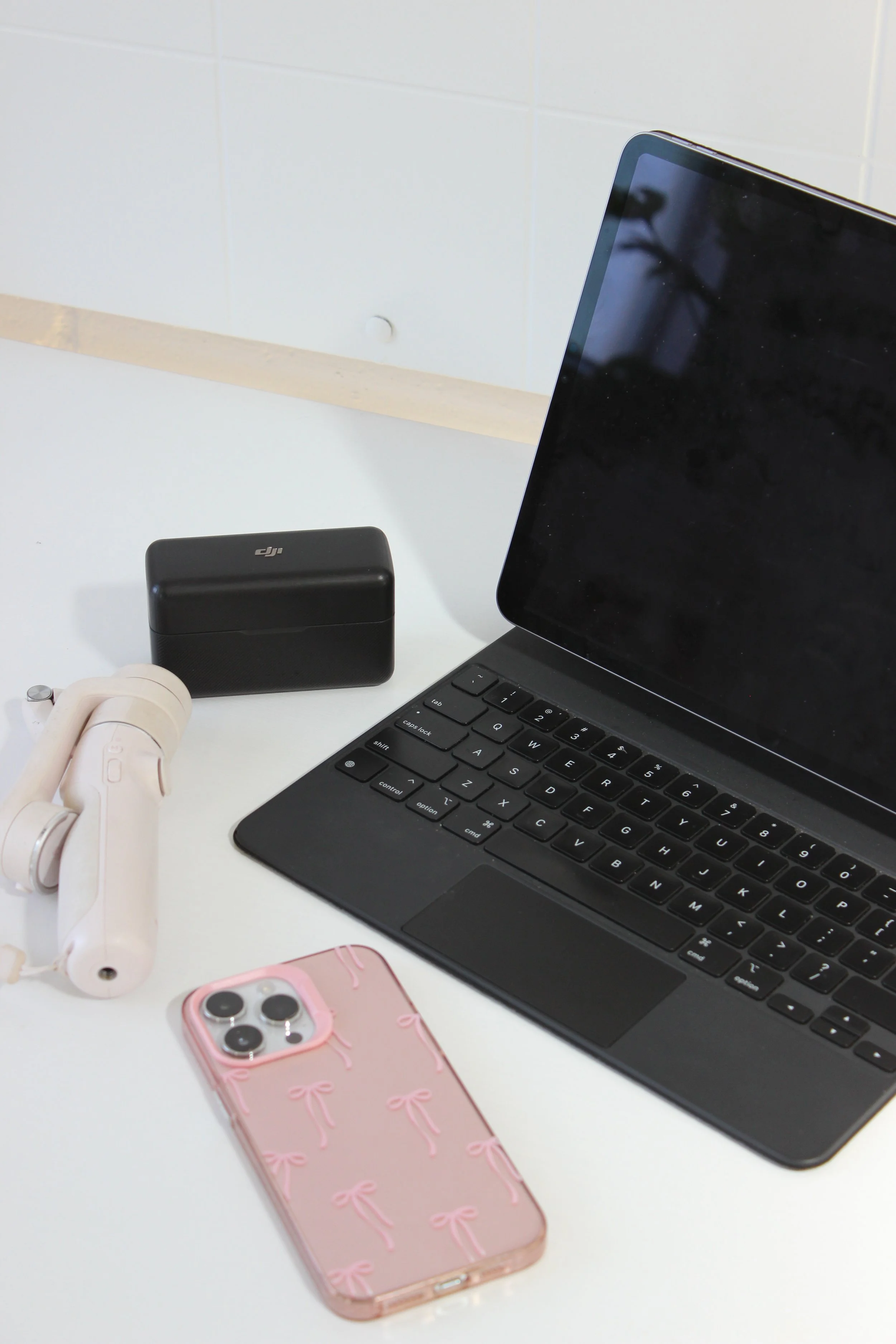 A laptop, pink phone with bows, white headphones, black case, and a small black drone controller on a white table.