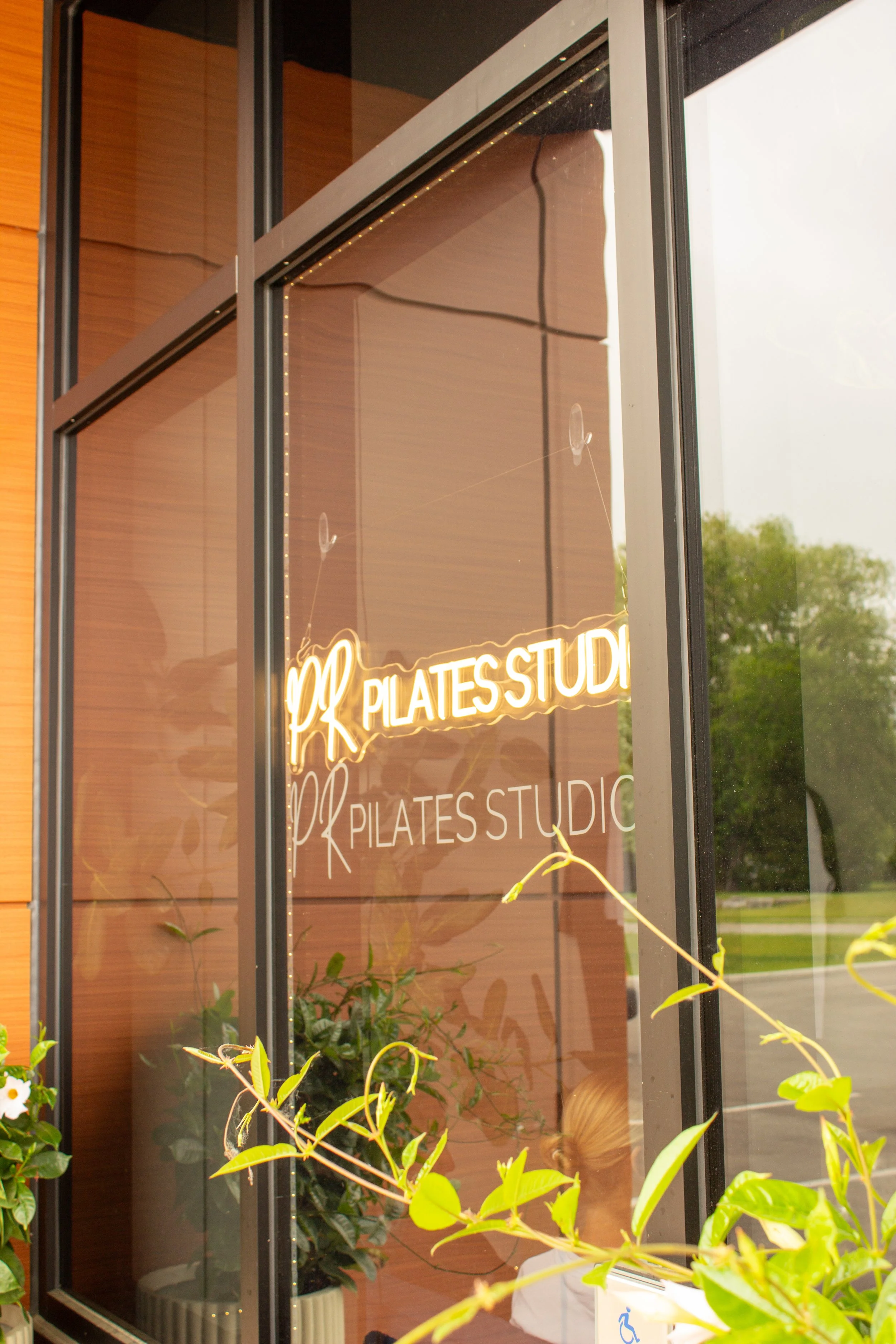 A glass window with a neon sign that reads 'Pilates Studio', surrounded by greenery and interior plants.