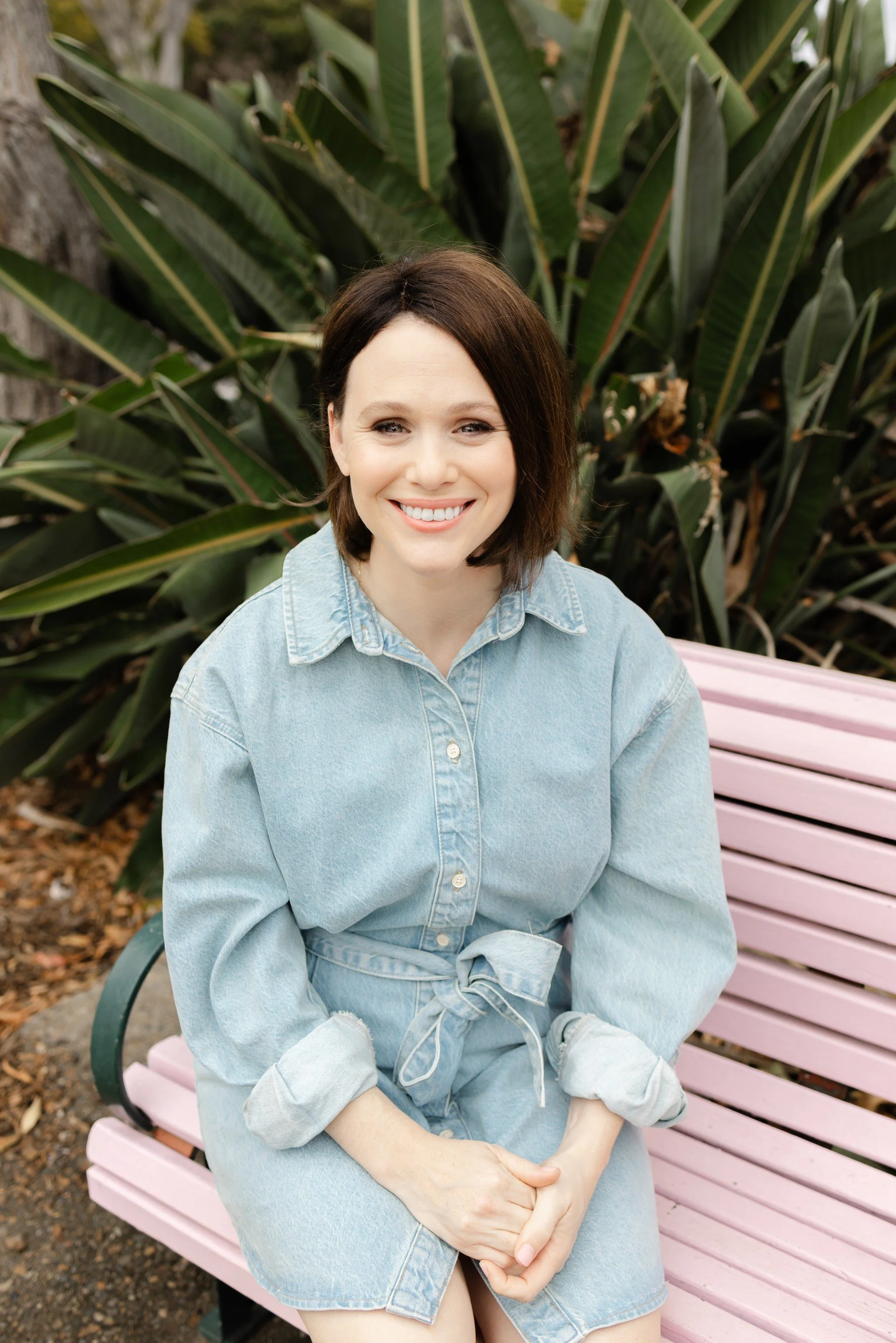Professional headshot of a female copywriter smiling outdoors, wearing a light denim dress, with greenery in the background.