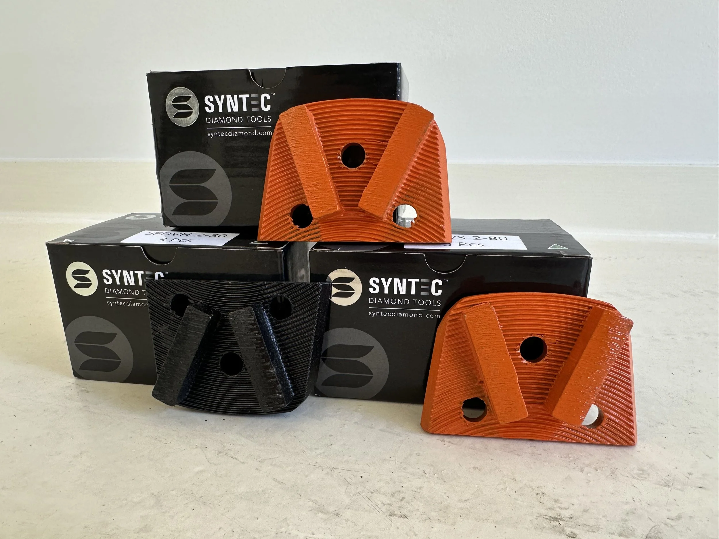 Three diamond grinding tool pads, two orange and one black, placed in front of Jack black boxes labeled 'SYNTÉC Diamond Tools'.