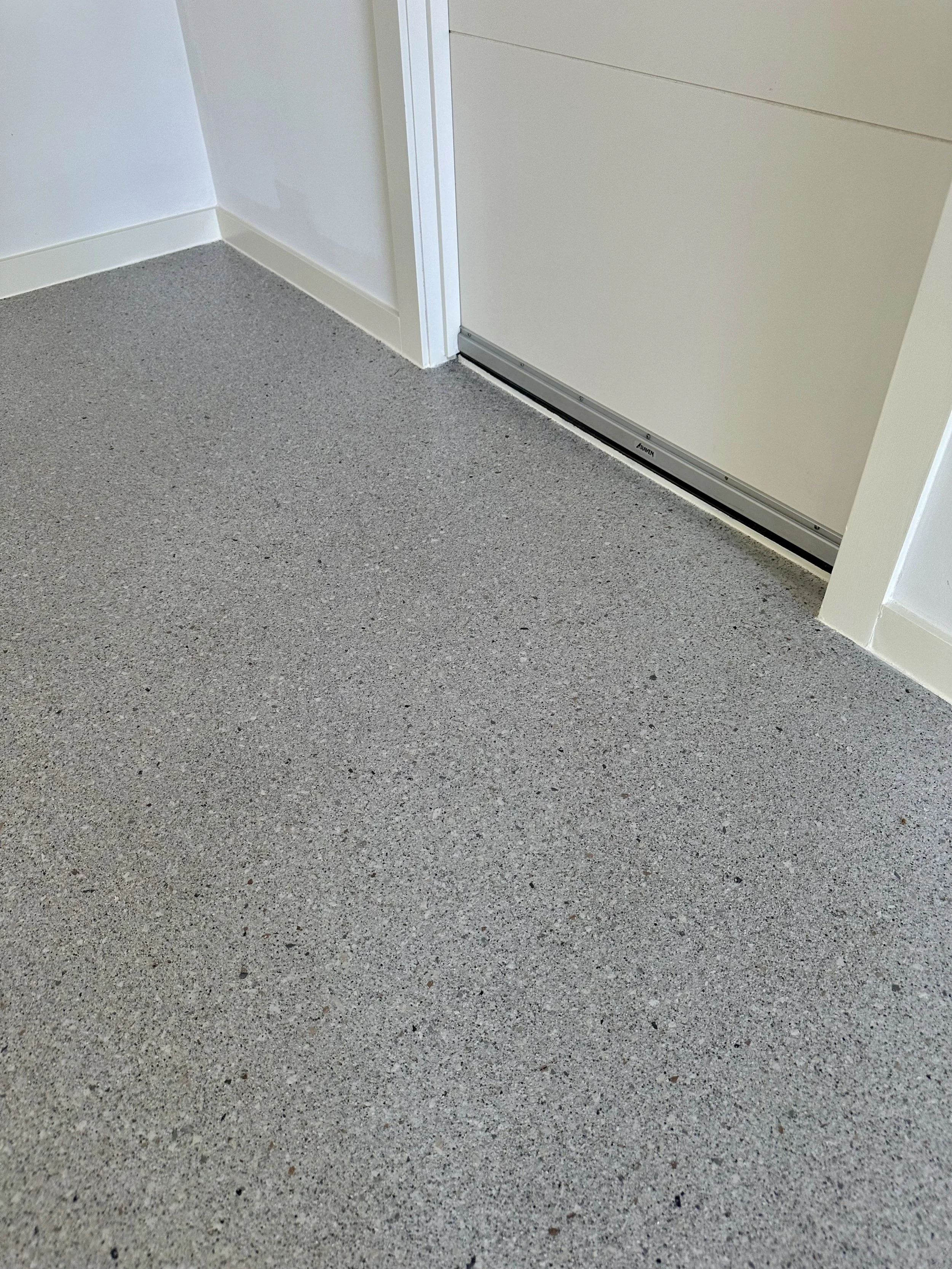 Close-up of a speckled gray rubber or vinyl floor with a white wall, baseboard, and a white door with a metallic door sweep.