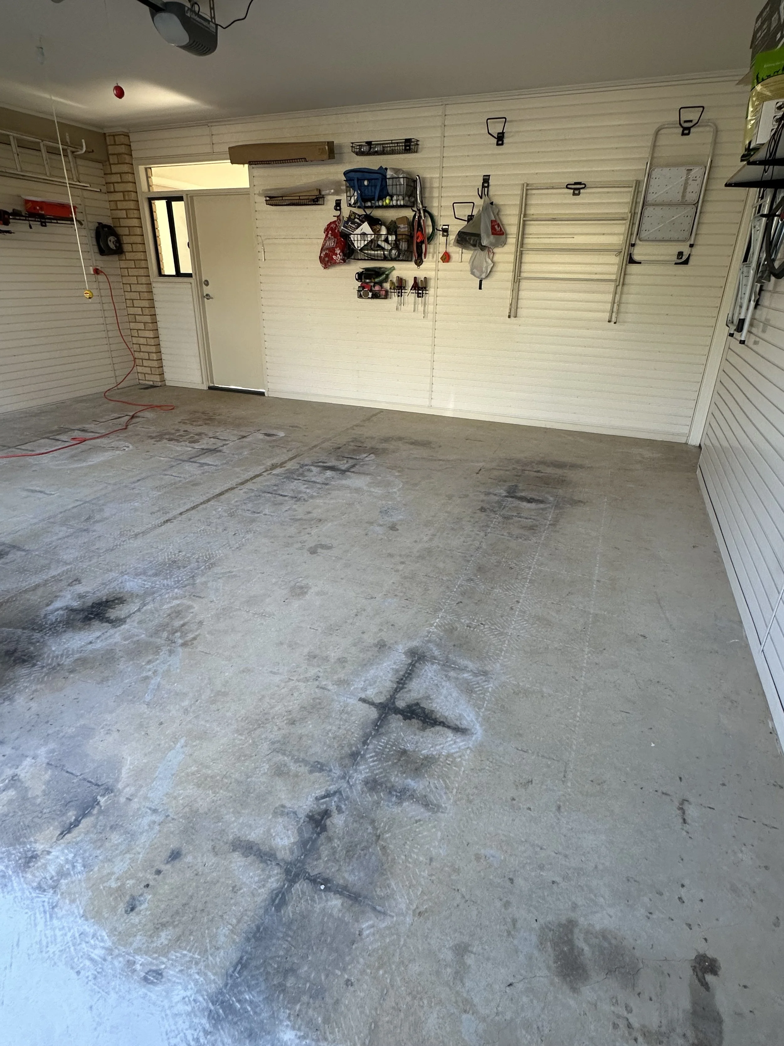 Empty garage with some black stains on the concrete floor, wall-mounted shelves with tools and supplies, and an open door leading outside.