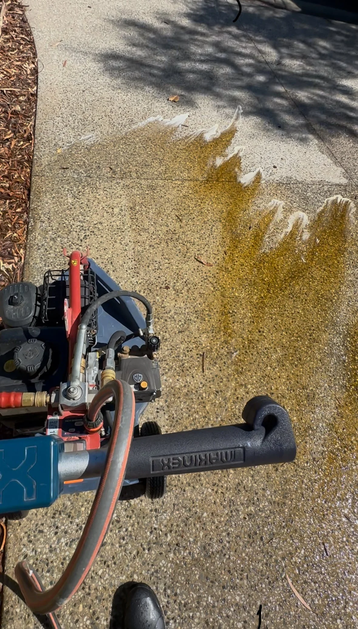 Close-up of a power washer spraying water onto a concrete sidewalk, with a shadow of tree branches overhead.