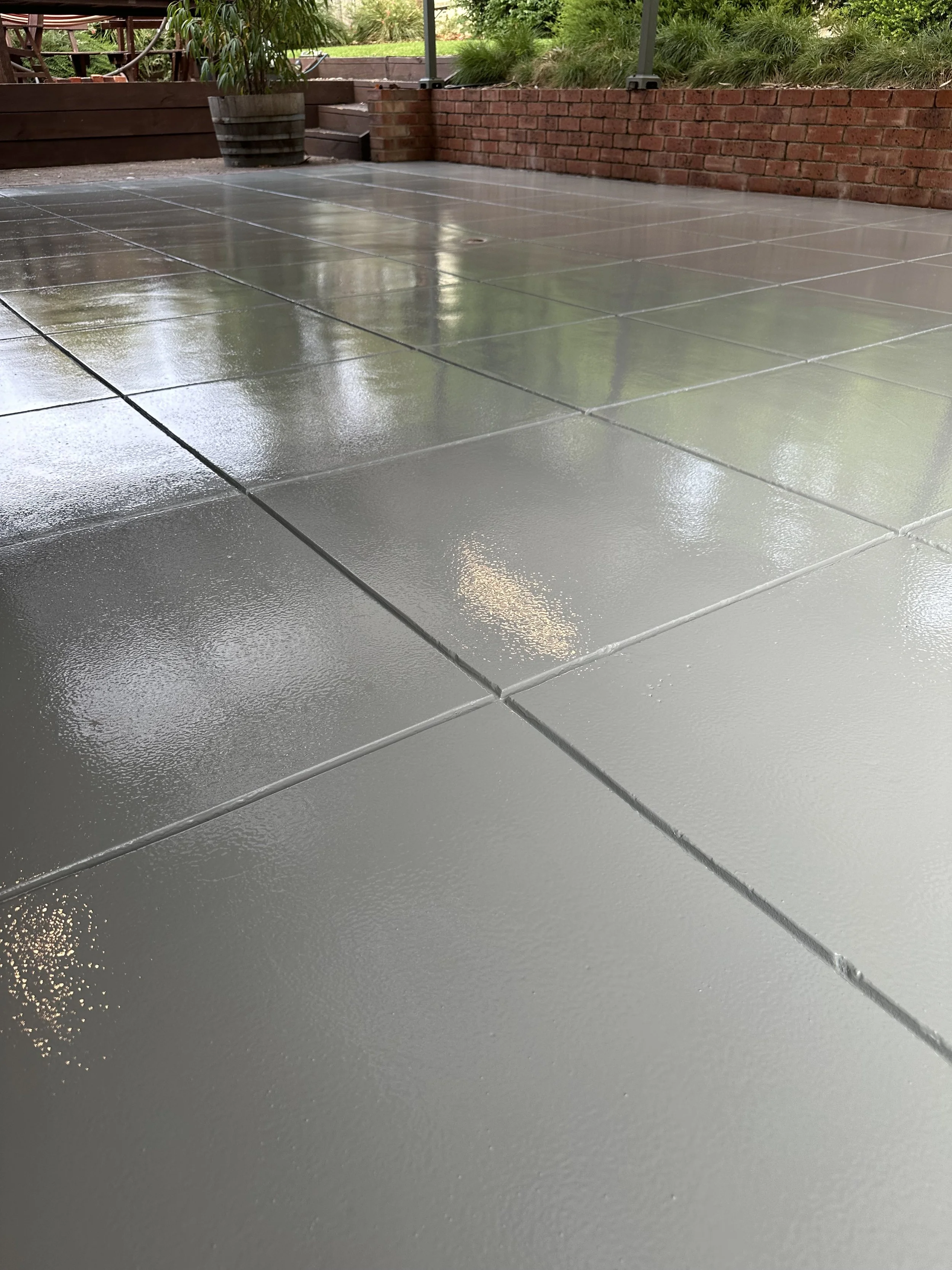 Wet, shiny, gray tiles on a patio with a brick wall and greenery in the background.