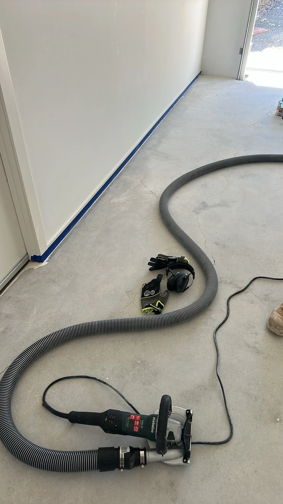 A concrete floor in a room with construction tools including a shop vacuum, gloves, ear protection, and a power tool for drywall finishing, with a partially finished wall and door in the background.