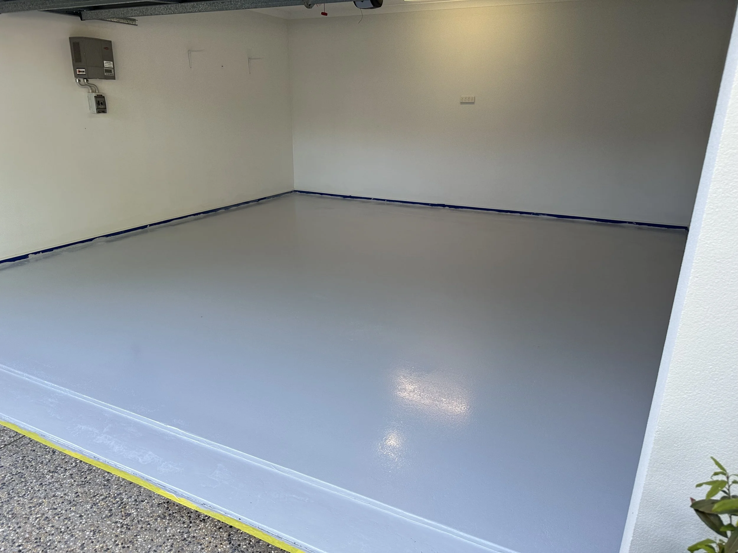 An empty enclosed parking space with a freshly painted light gray floor, white walls, and some electrical and utility panels on the wall.