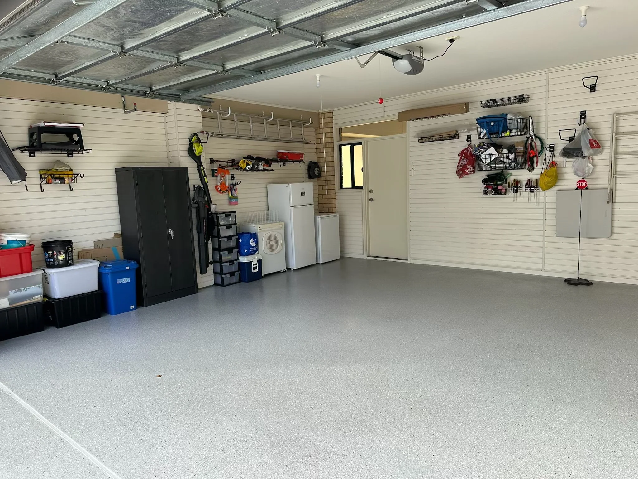 Clean, organized garage with tools, storage shelves, a black cabinet, and a washer and dryer. The garage door is open, and various gardening and household items are neatly stored.