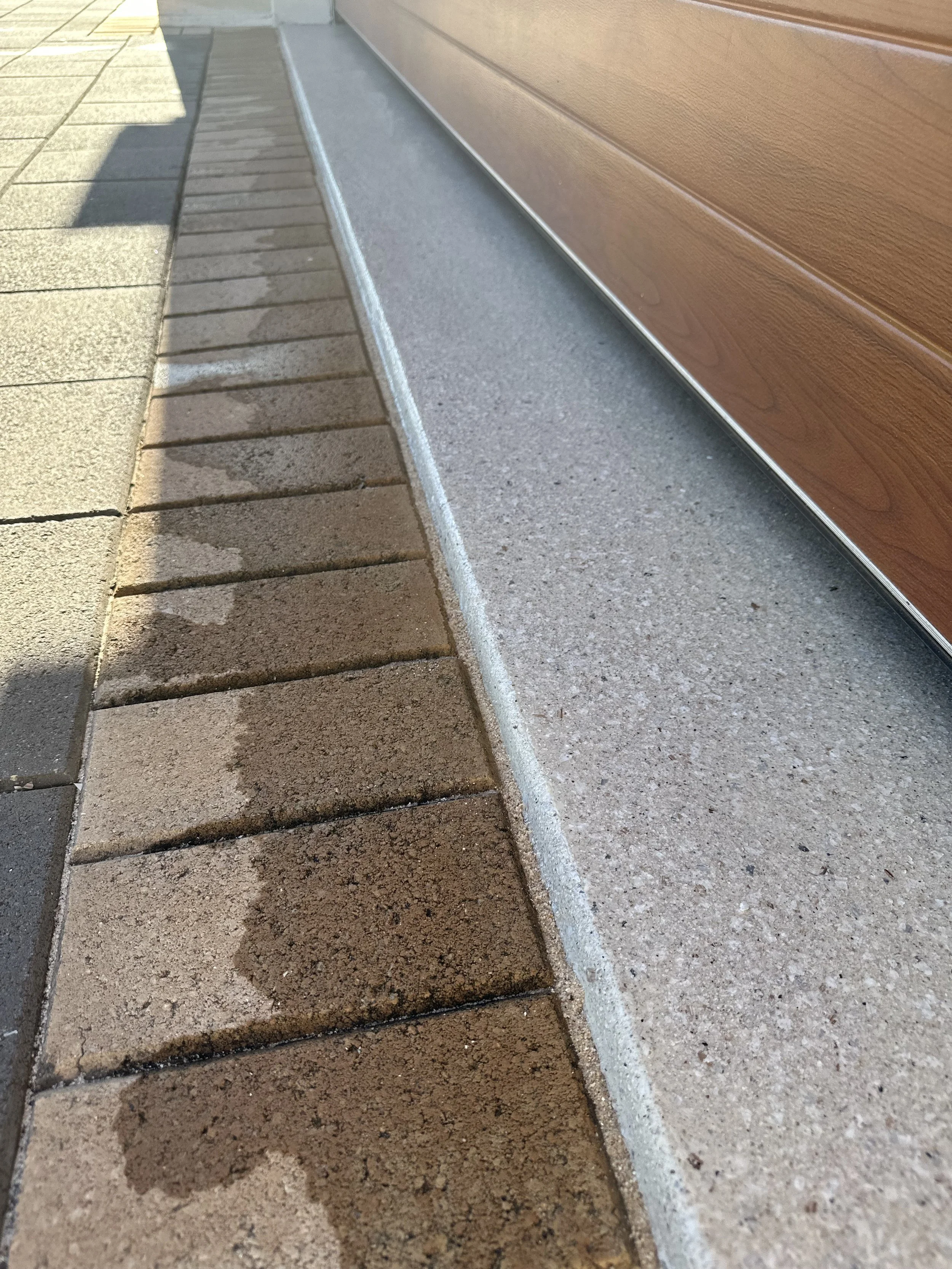 Close-up of a concrete and brick sidewalk next to a brown wooden house, showing a freshly poured concrete strip between the bricks and the house.