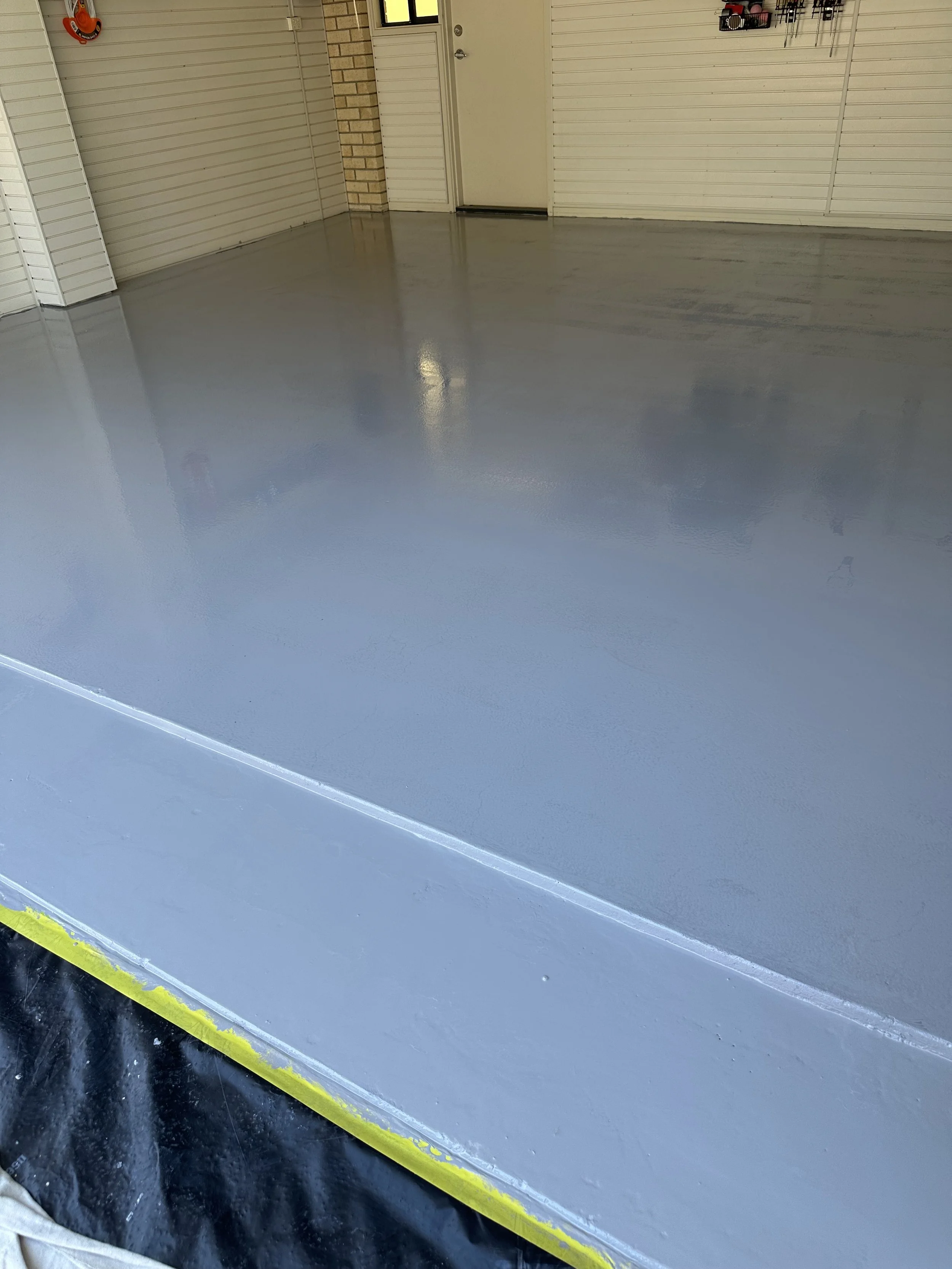 Freshly painted gray epoxy floor in a garage with beige walls, a door, and some tools hanging on the wall.