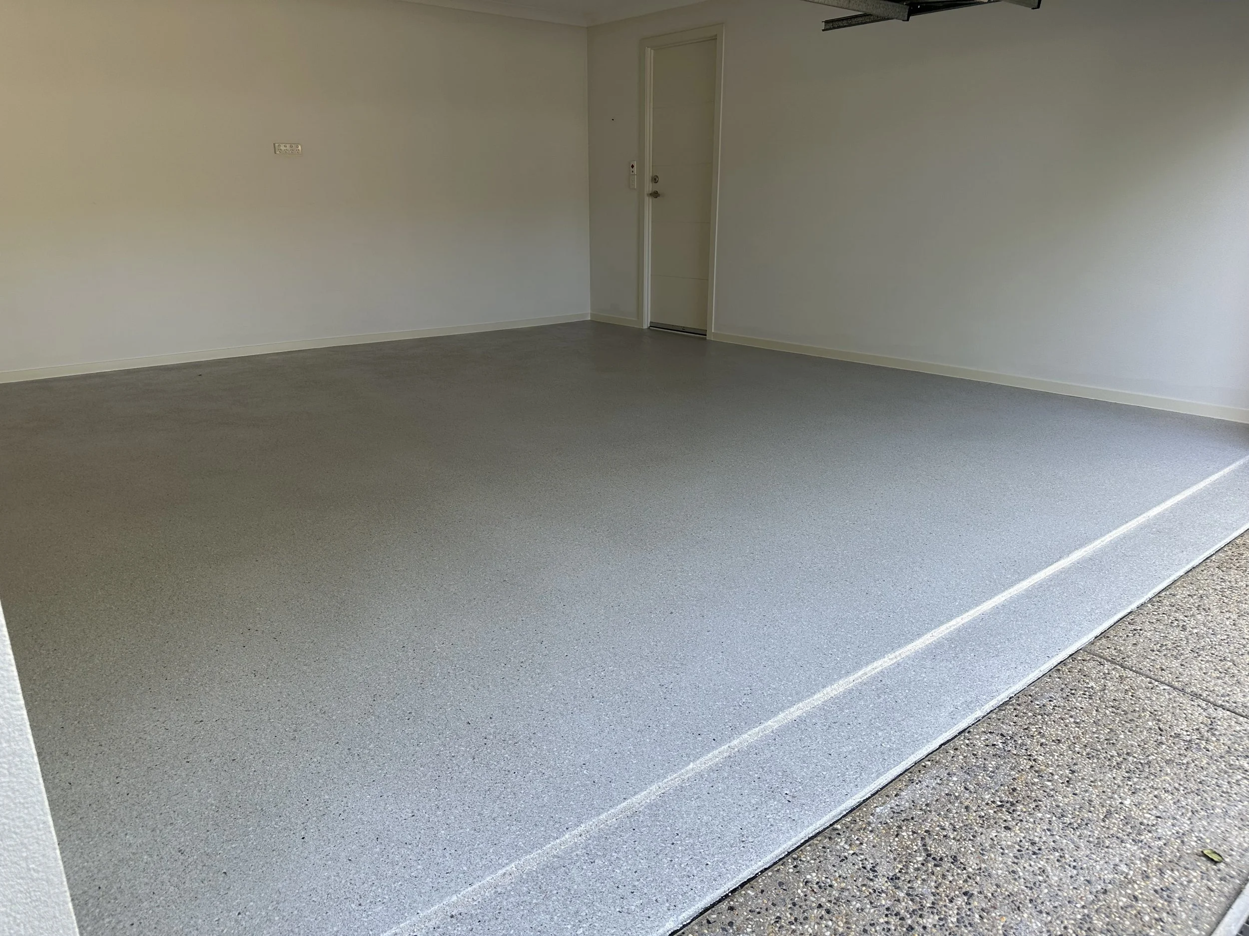 Empty garage with light gray textured floor, white walls, closed white door, and electrical outlets.