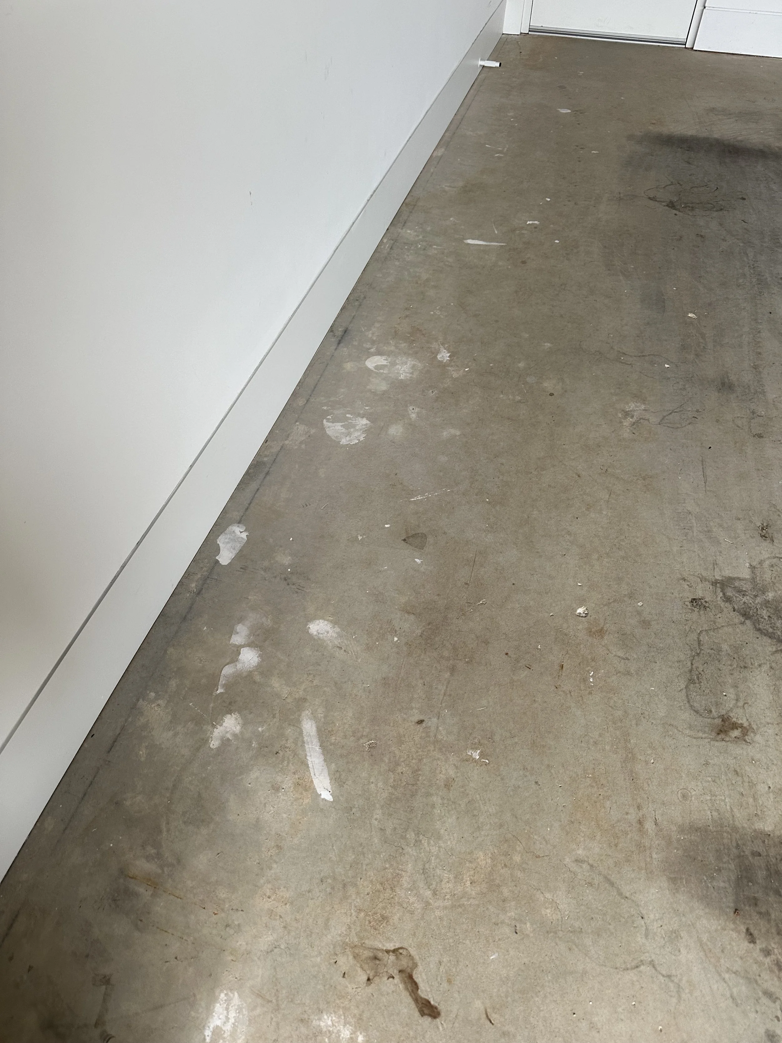 Concrete floor with white paint splatters and scuff marks next to a white wall and baseboard, near a closed door.