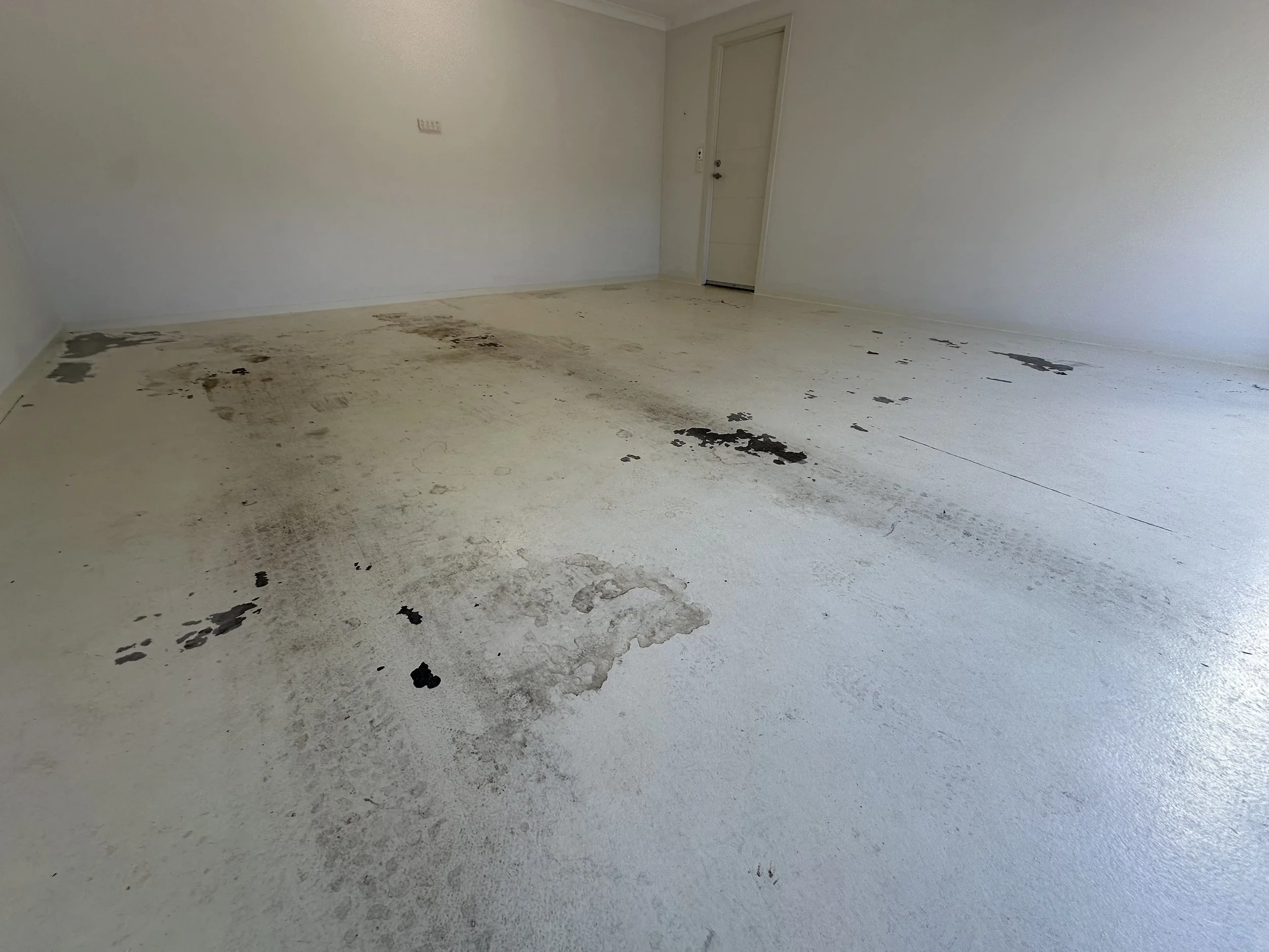 Room with stained and dirty white floor, with dark spots and patches of black and gray dirt or mold near the wall and around the door.