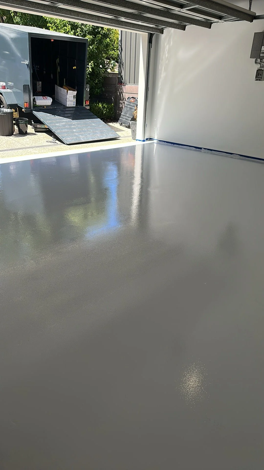 Freshly painted concrete floor in a garage with a partially open garage door, revealing outdoor equipment and a trailer.