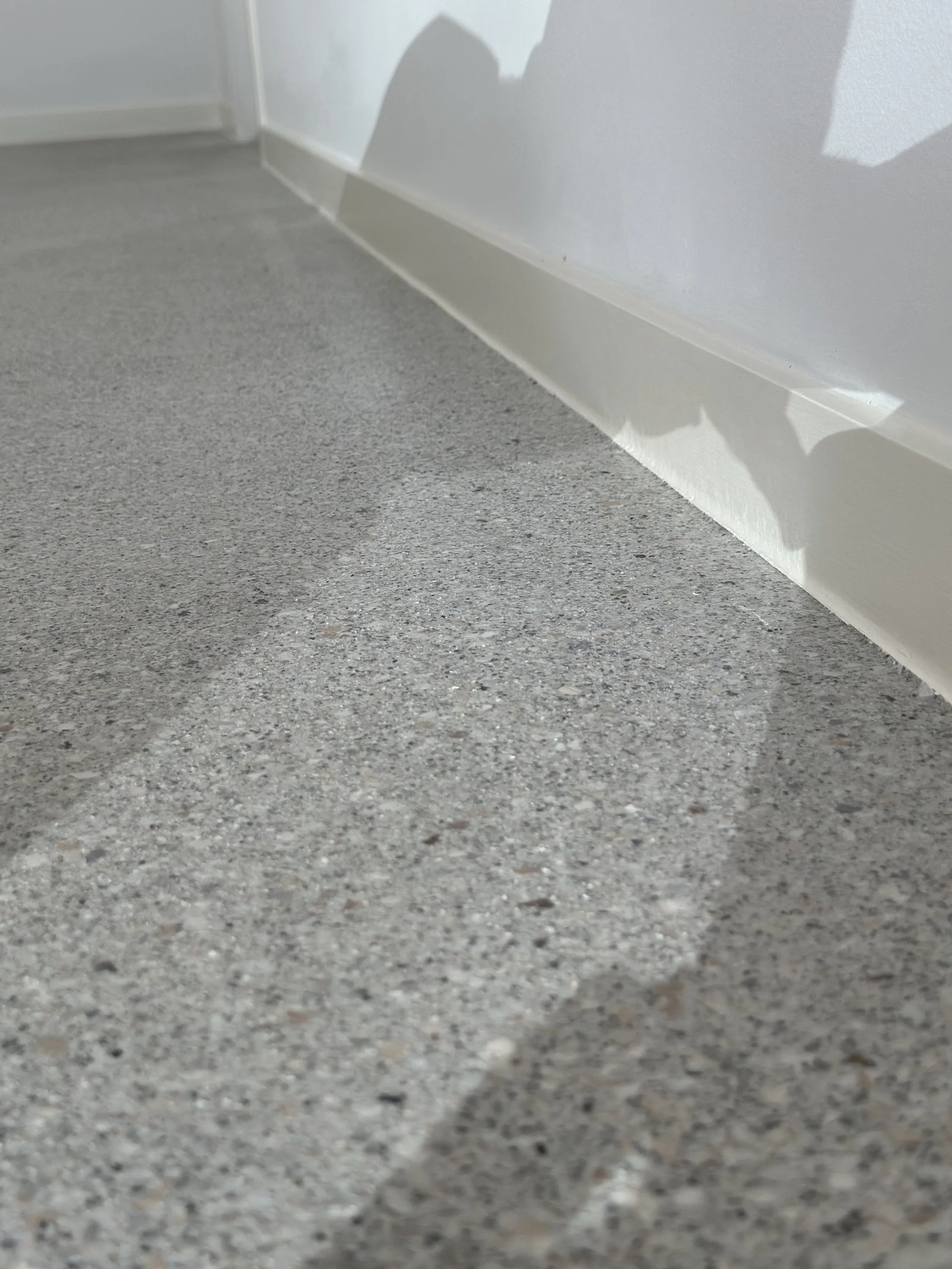 Close-up of a textured gray carpeted floor near a white baseboard and white wall with a shadow.