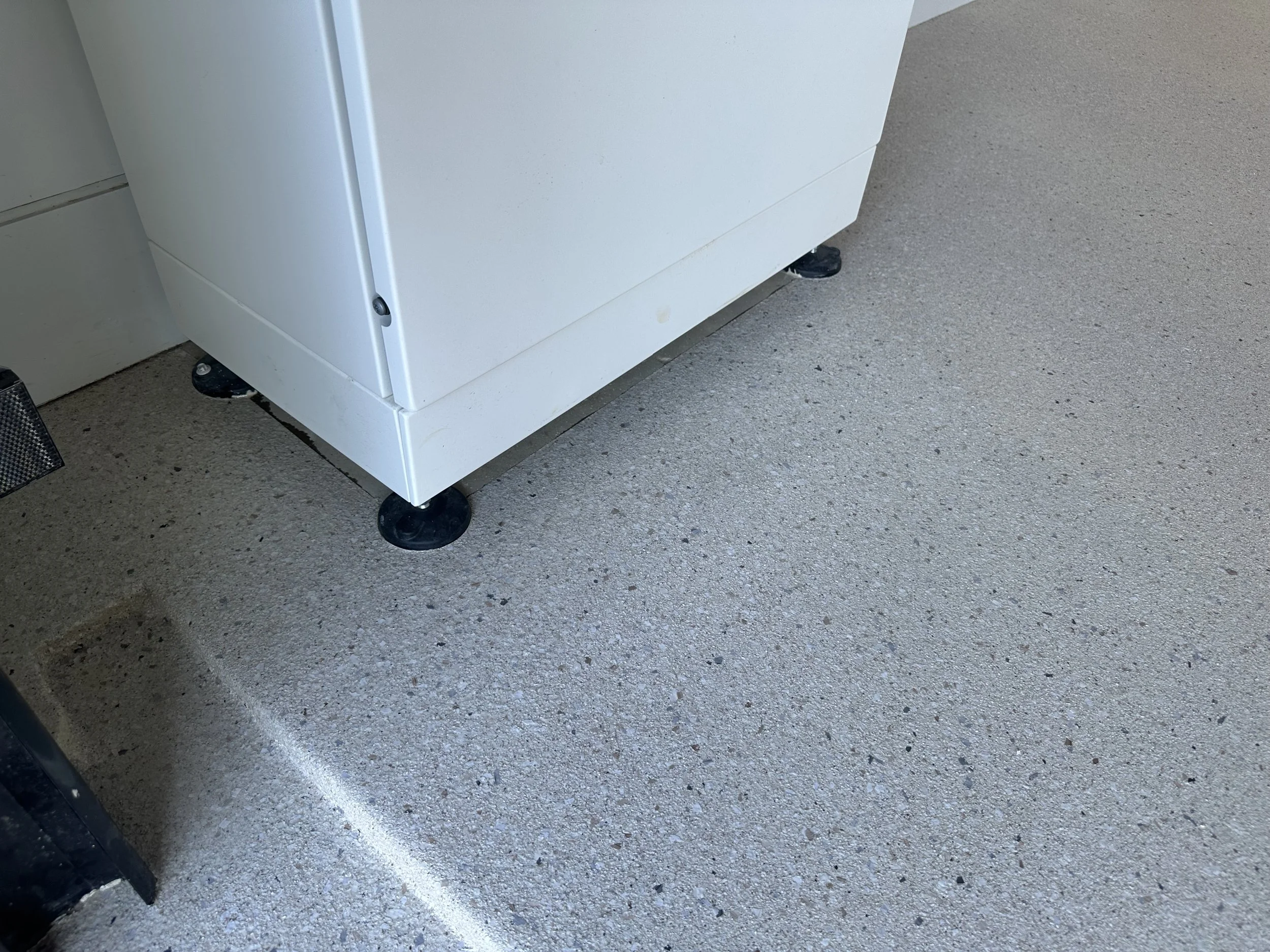 Close-up of the bottom corner of a white appliance on four black caster wheels, on a speckled gray concrete floor.
