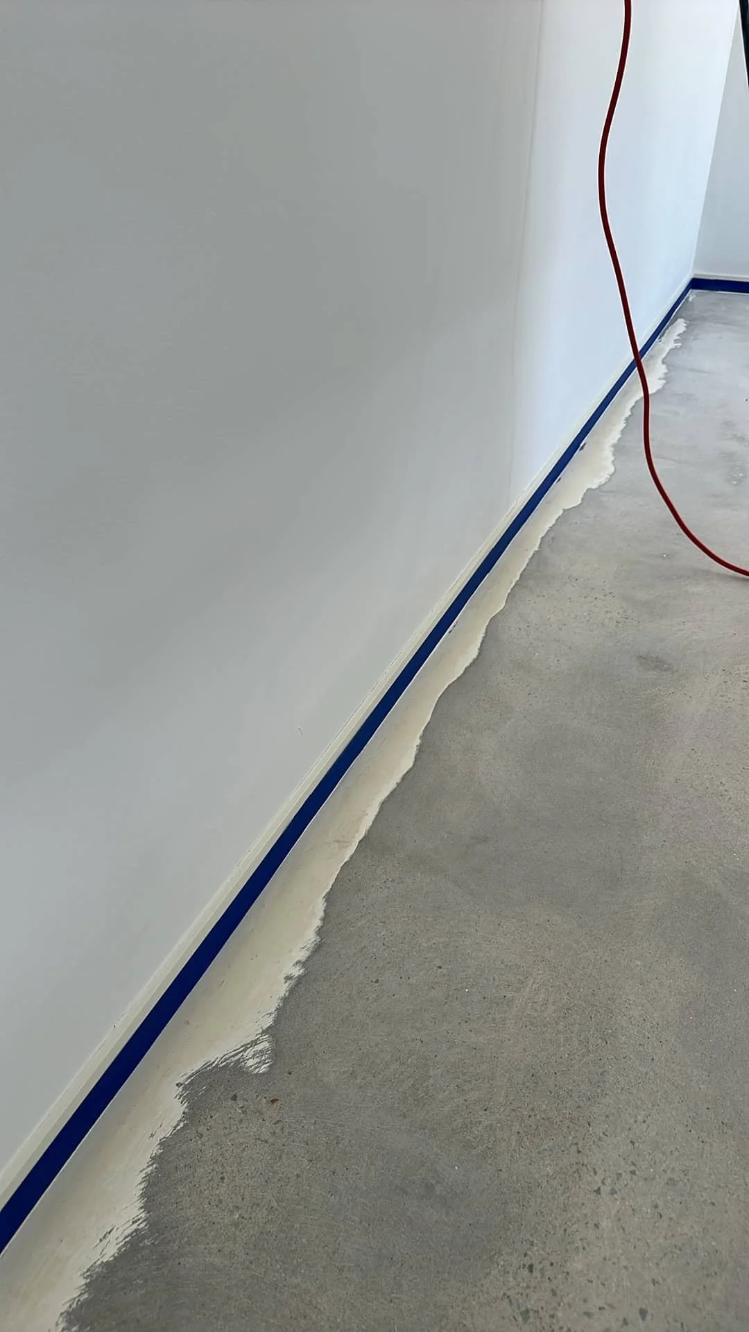 Construction scene showing tape and sealant along the base of a white wall adjacent to a concrete floor.