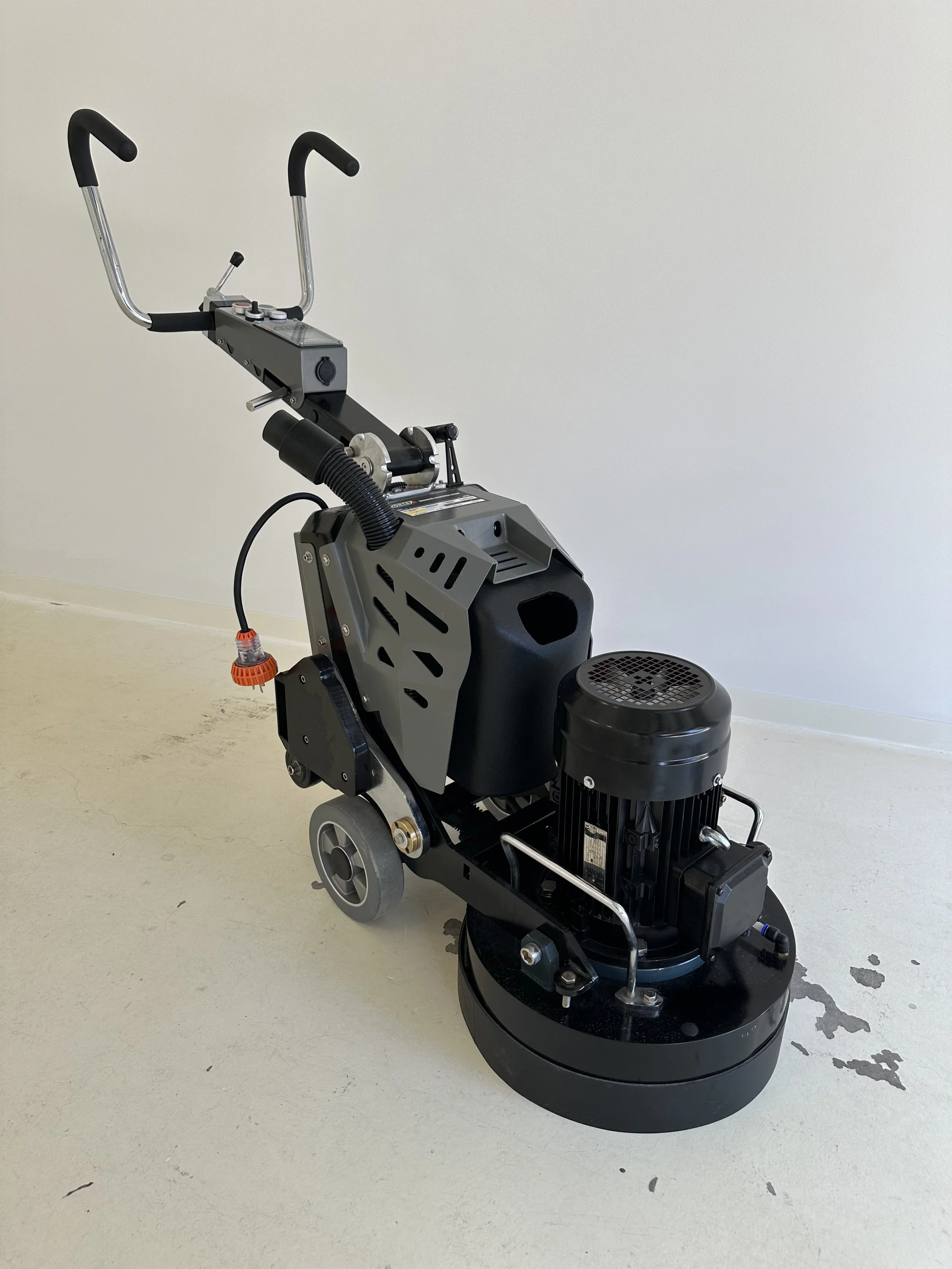 Floor polishing machine with a black circular base, motor, and handle, positioned on a beige floor against a plain wall.