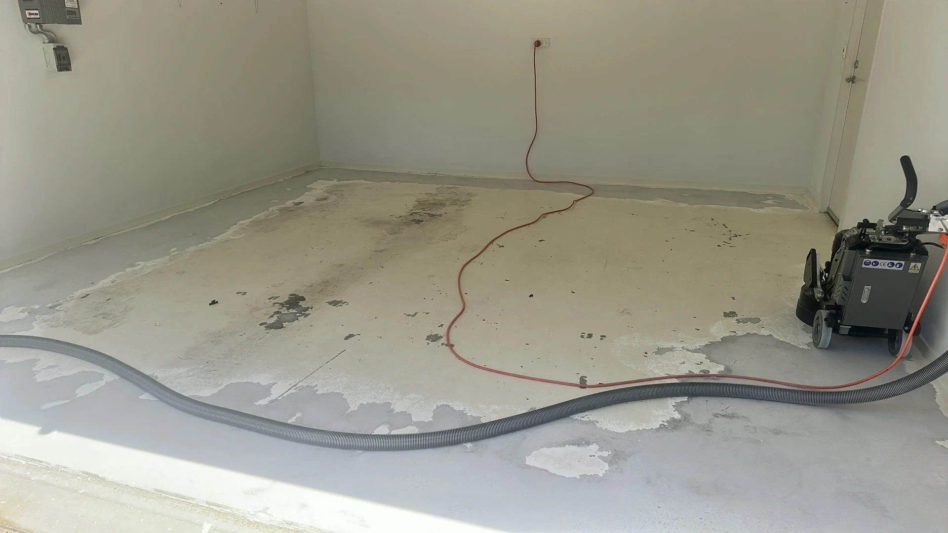 Empty room with peeling paint on the floor, a hose and a machine possibly for construction or cleaning, and exposed electrical outlets.