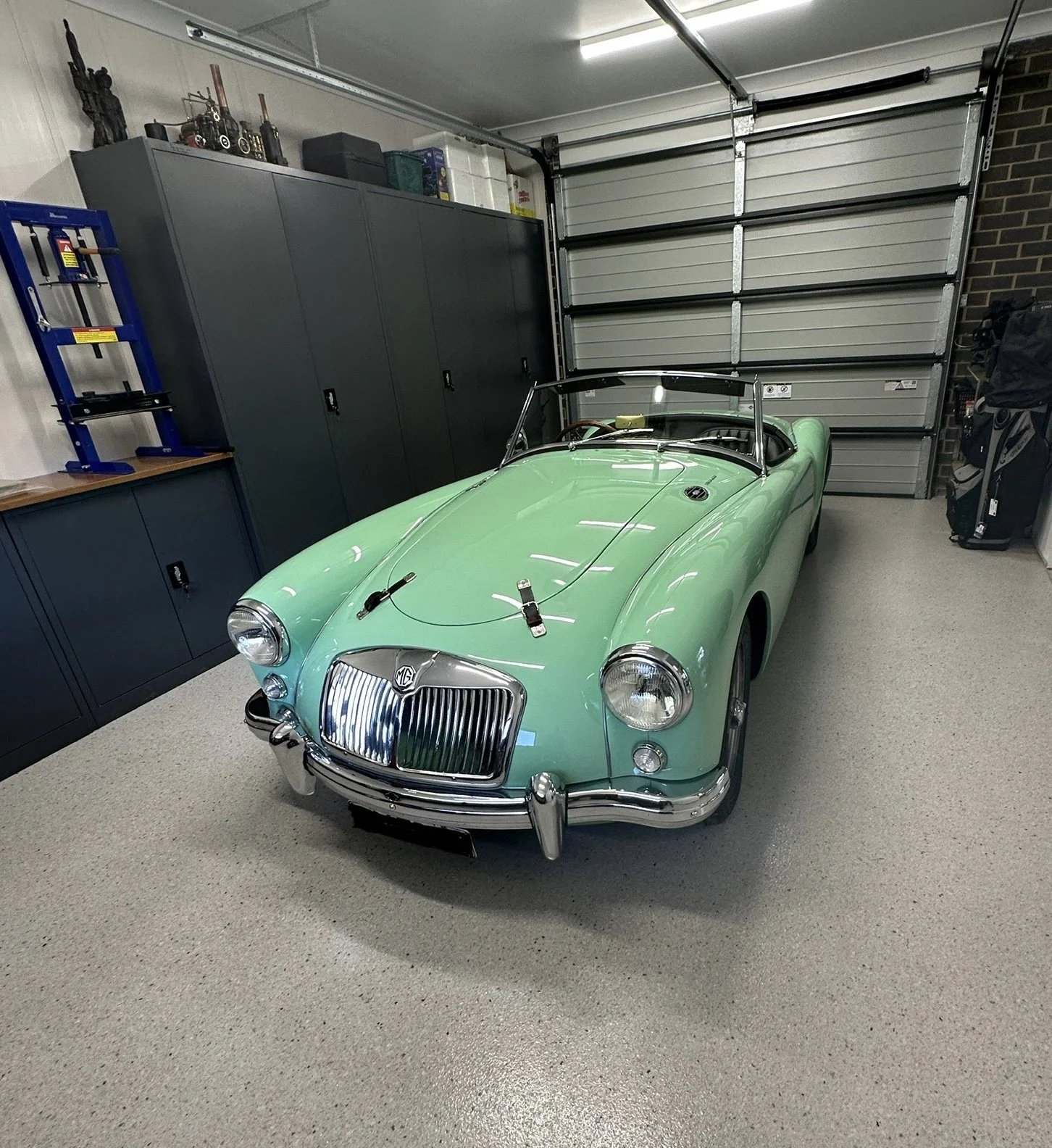 A vintage mint green MG convertible sports car parked in a garage with gray cabinets and a silver garage door.