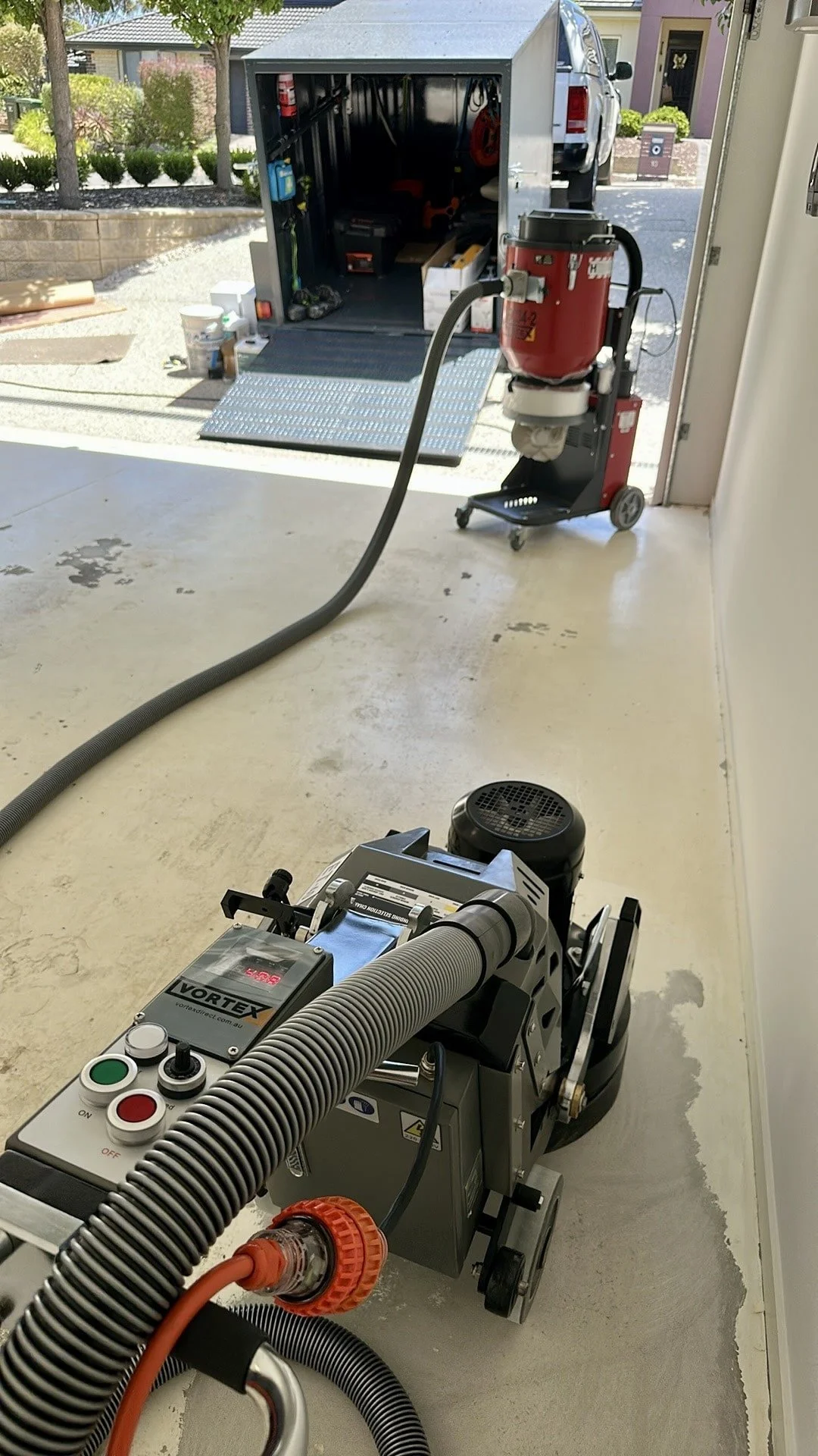 Portable industrial vacuum cleaner with a flexible hose connected, in a garage. In the background, a vehicle with an open trailer filled with tools and equipment.