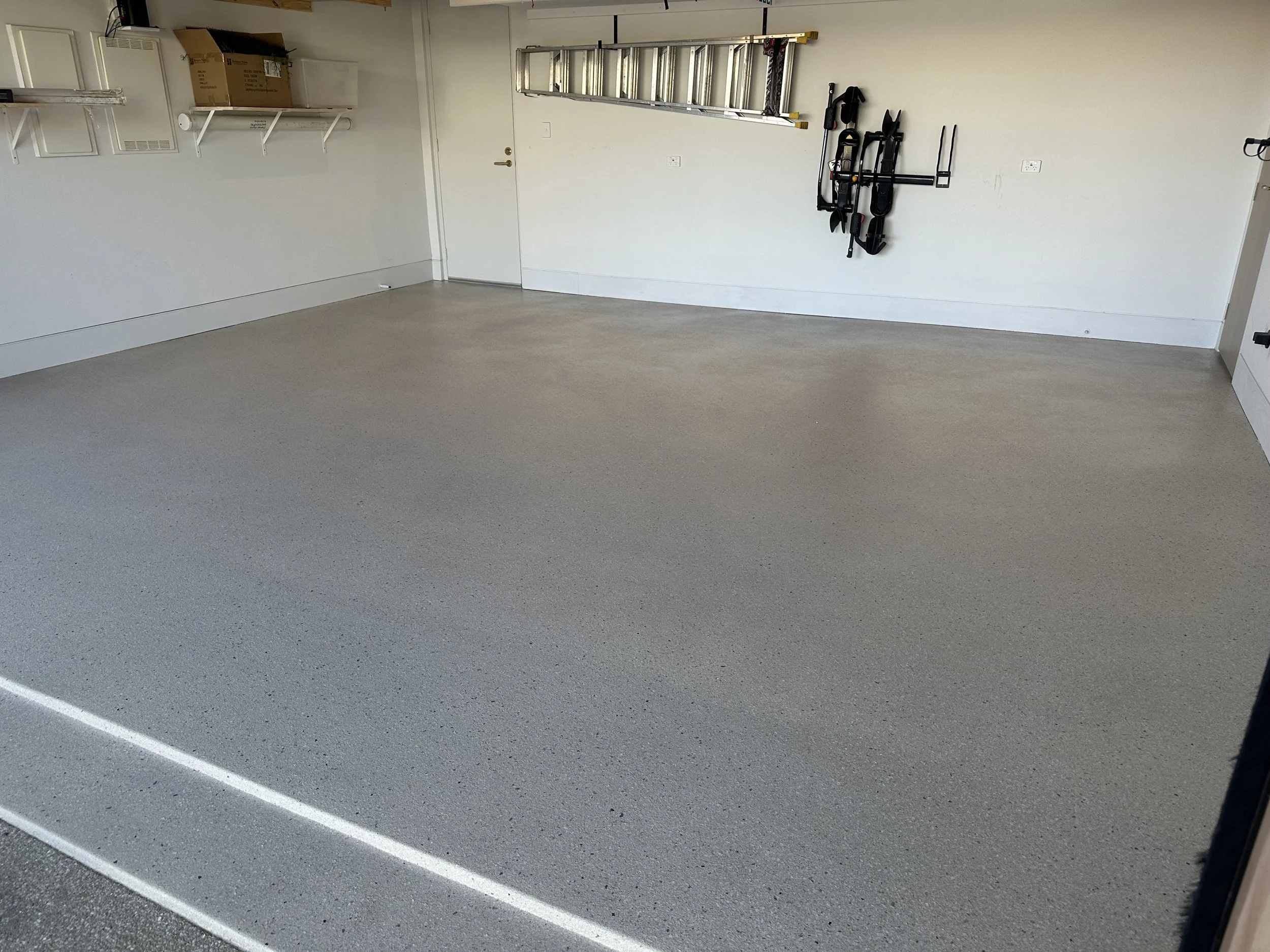 Empty garage with a clean, speckled gray floor, white walls, shelves, a ladder, and storage items on the walls.