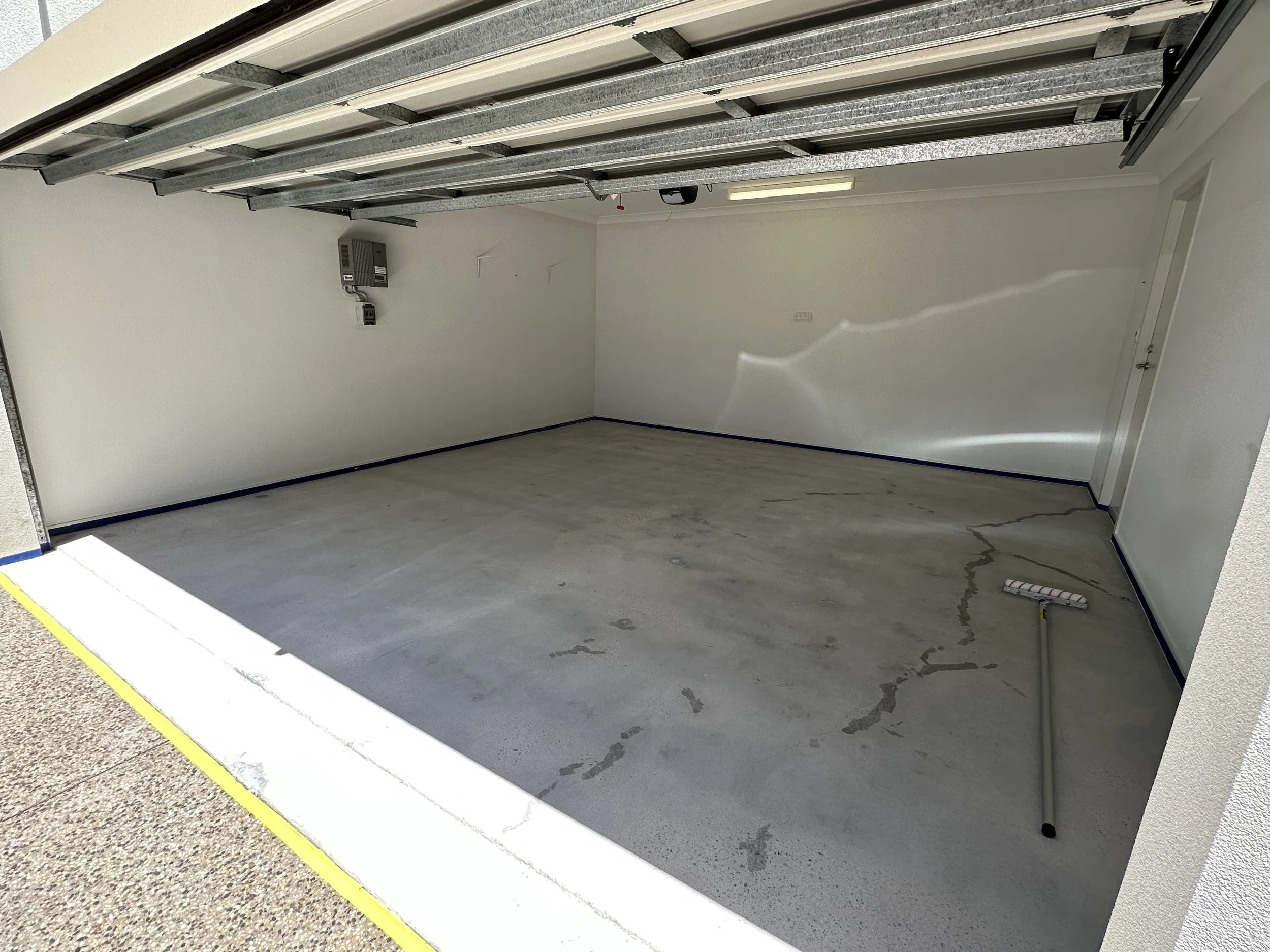 Empty residential garage with open door, concrete floor, white walls, and a broom leaning against the wall.