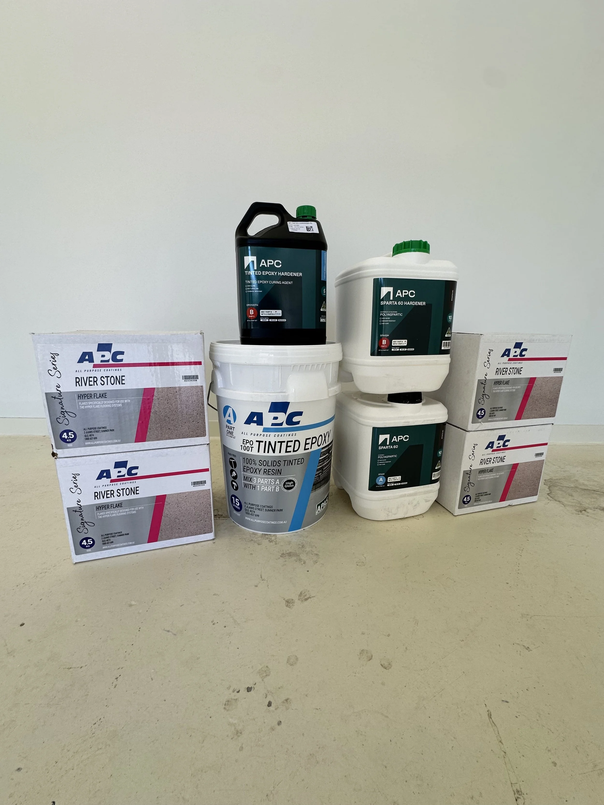 Various containers and boxes of APC brand epoxy, hardener, primer, and coating products on a beige surface.
