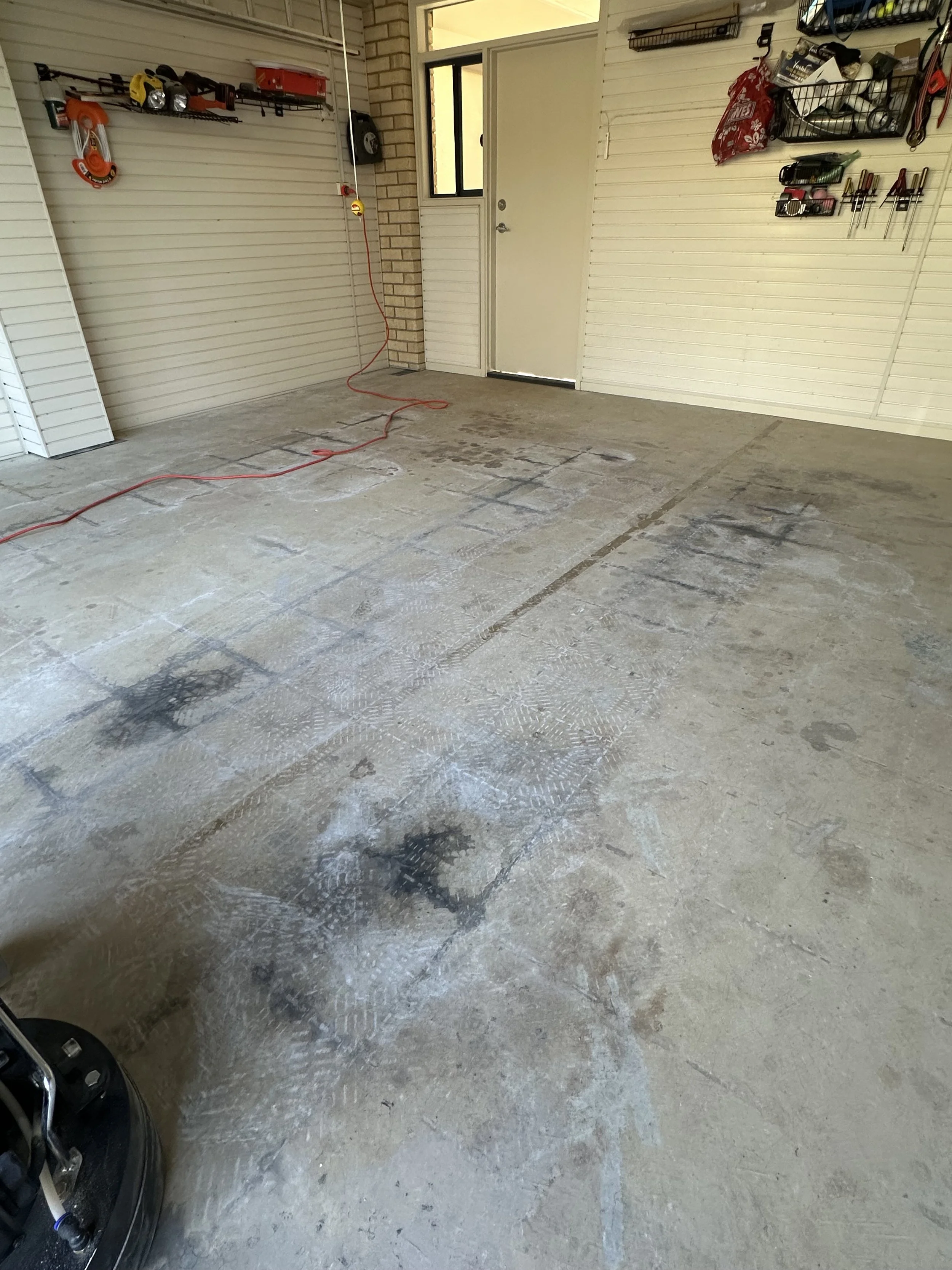 Empty garage with a concrete floor, some dark stains and footprints, tools and supplies on the walls, and a red extension cord on the floor.