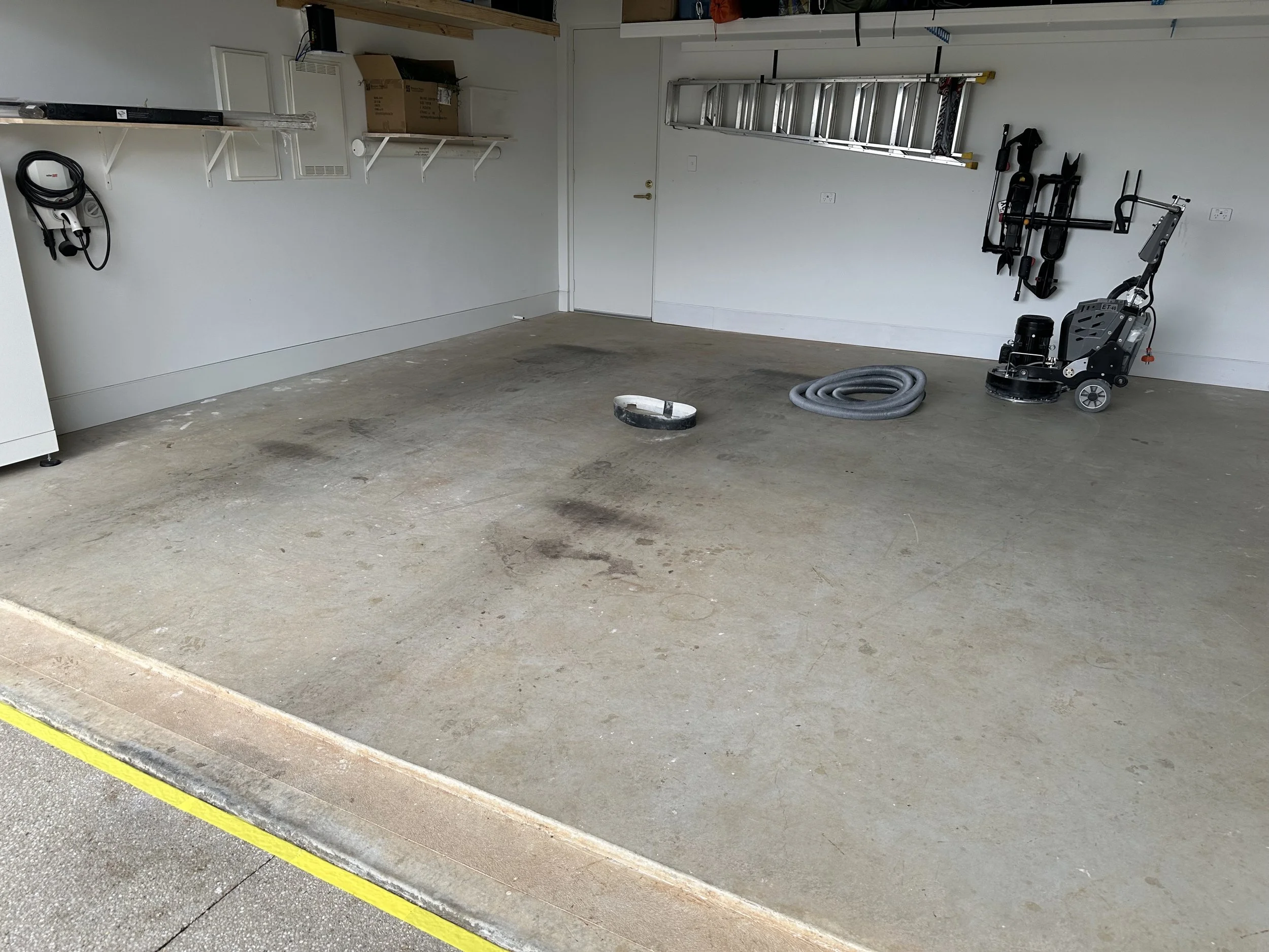 Empty garage with cleaning equipment, including a vacuum cleaner, a coiled hose, and wall-mounted tools, with some stains on the concrete floor.