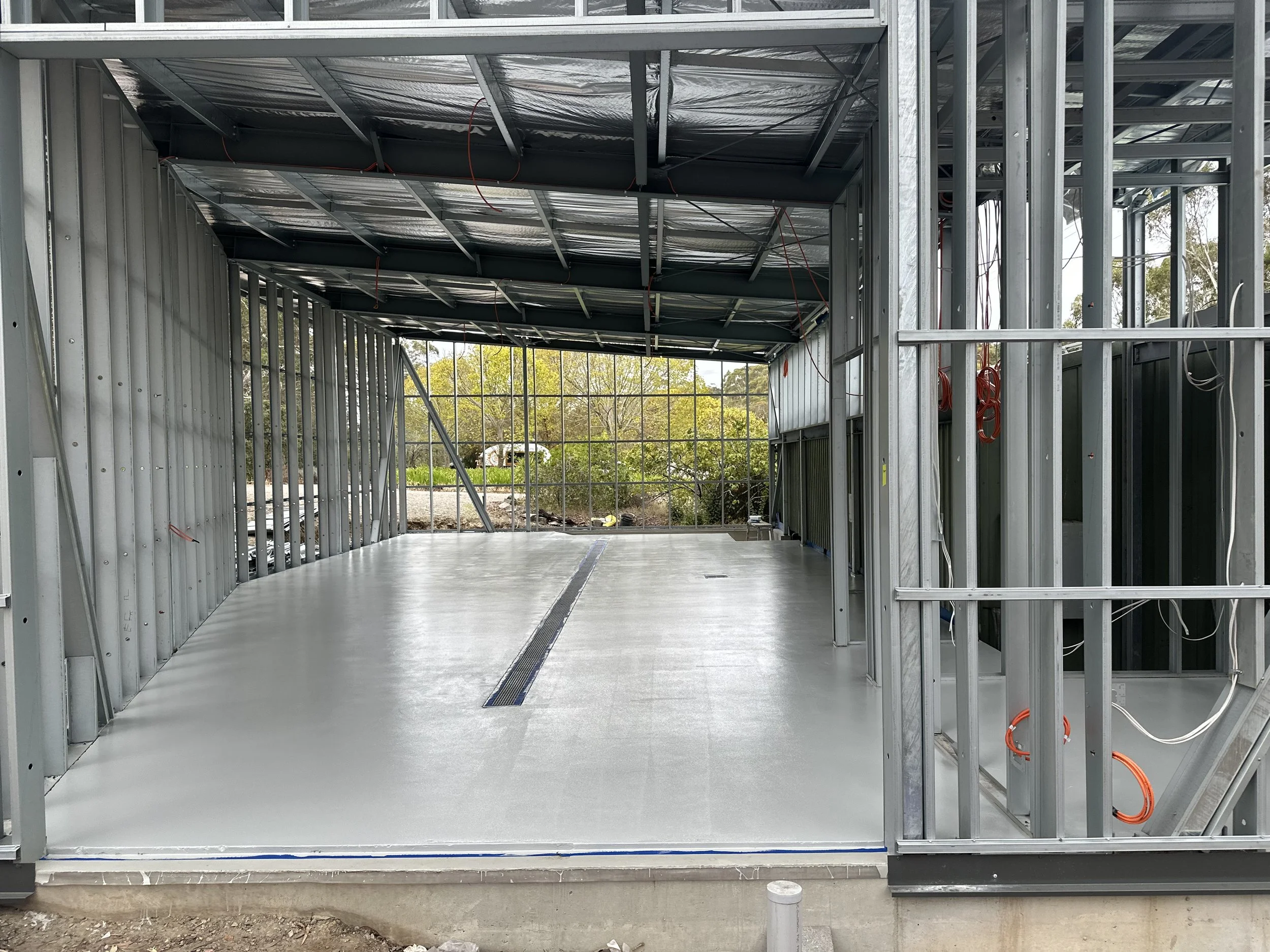 Steel-framed building under construction with a concrete floor and visible wiring, open on one side with trees visible in the background.