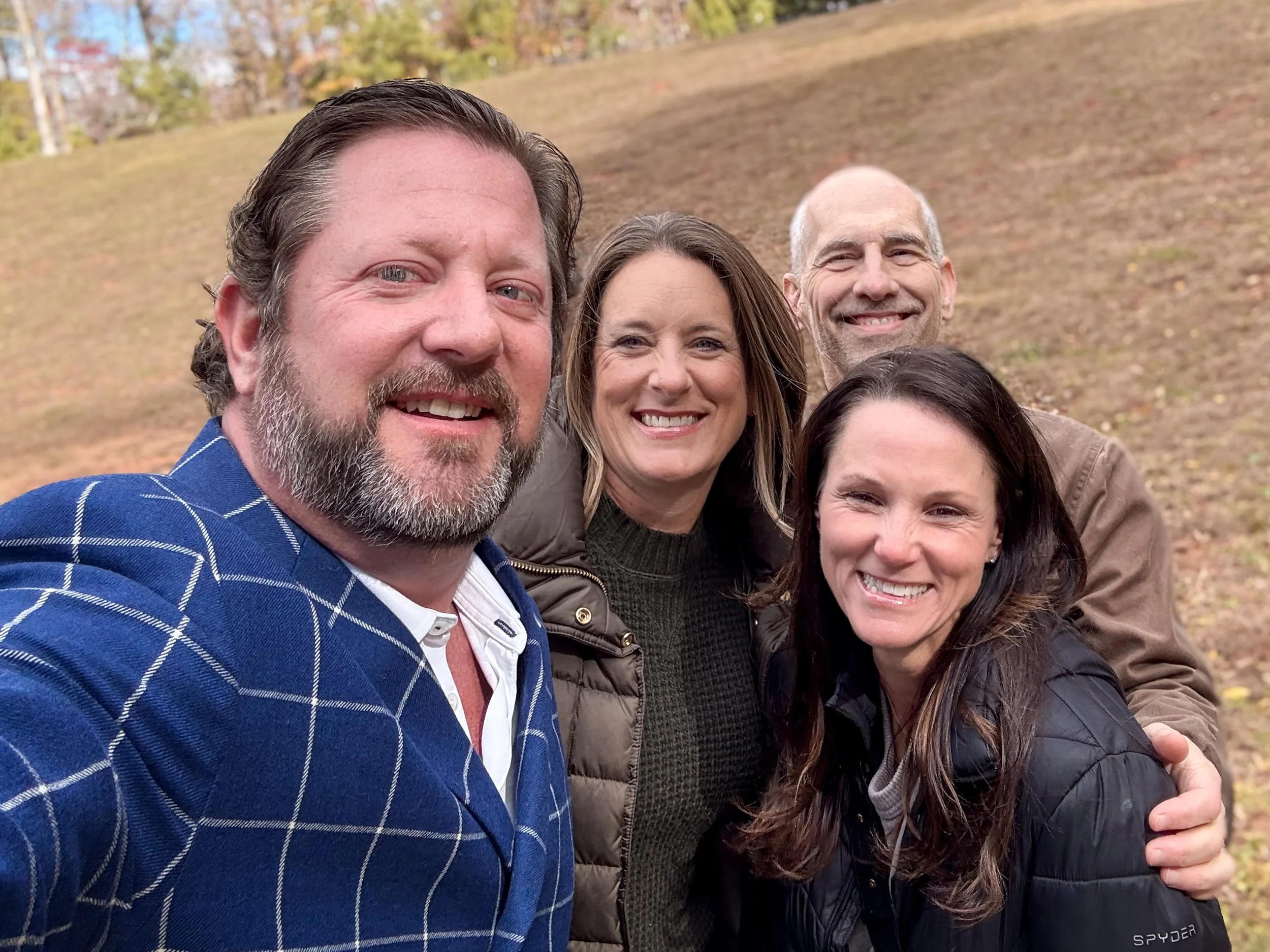A group of four adults, two men and two women, smiling and taking a selfie outdoors at a farm with autumn foliage in the background.