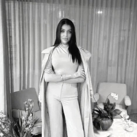 A young woman with long dark hair stands in a room with curtains, two chairs, plants, and a table with a teapot and a flower.