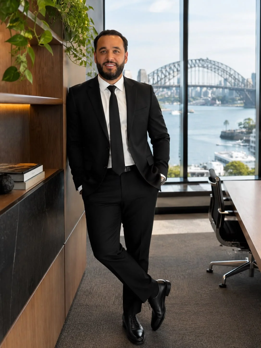 A man in a black suit with a white shirt and black tie standing indoors near a wooden shelf with books and plants, with a large window overlooking a river and a bridge in the background.